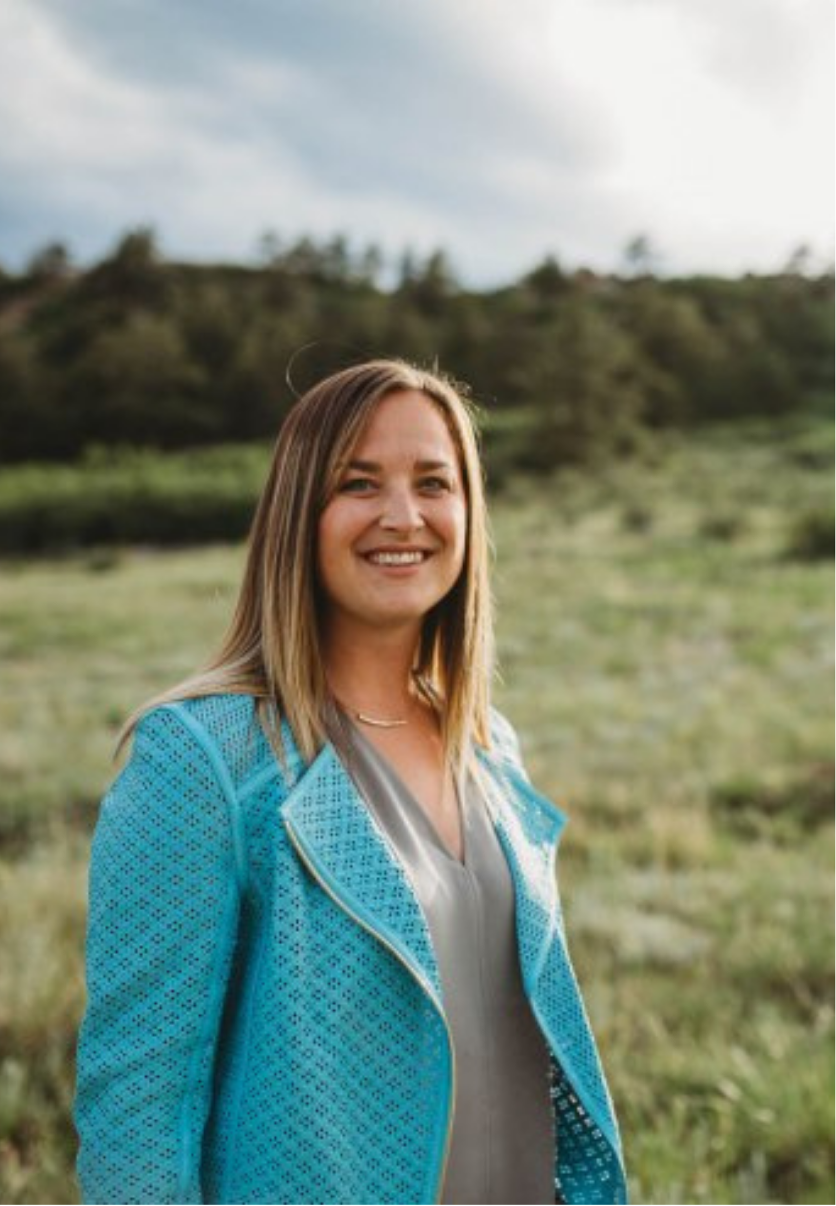 Kati Saul, therapist in Colorado Springs, with long blond hair smiling outdoors on a cloudy day, wearing a blue jacket over a beige top, with greenery and trees in the background.