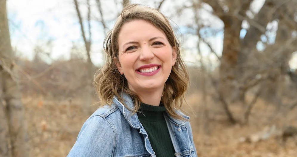 Jennie Radspinner, smiling with shoulder-length wavy brown hair, wearing a denim jacket, standing outdoors in a wooded area with leafless trees.