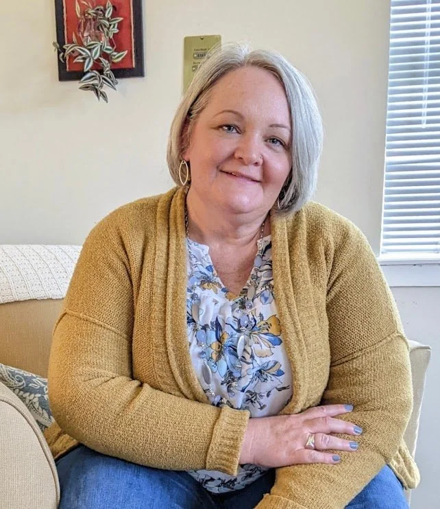 Lou Ann Marquez, therapist in Colorado Springs, with gray hair wearing a floral blouse and mustard yellow cardigan sitting on a beige couch in a room with a window and artwork on the wall.