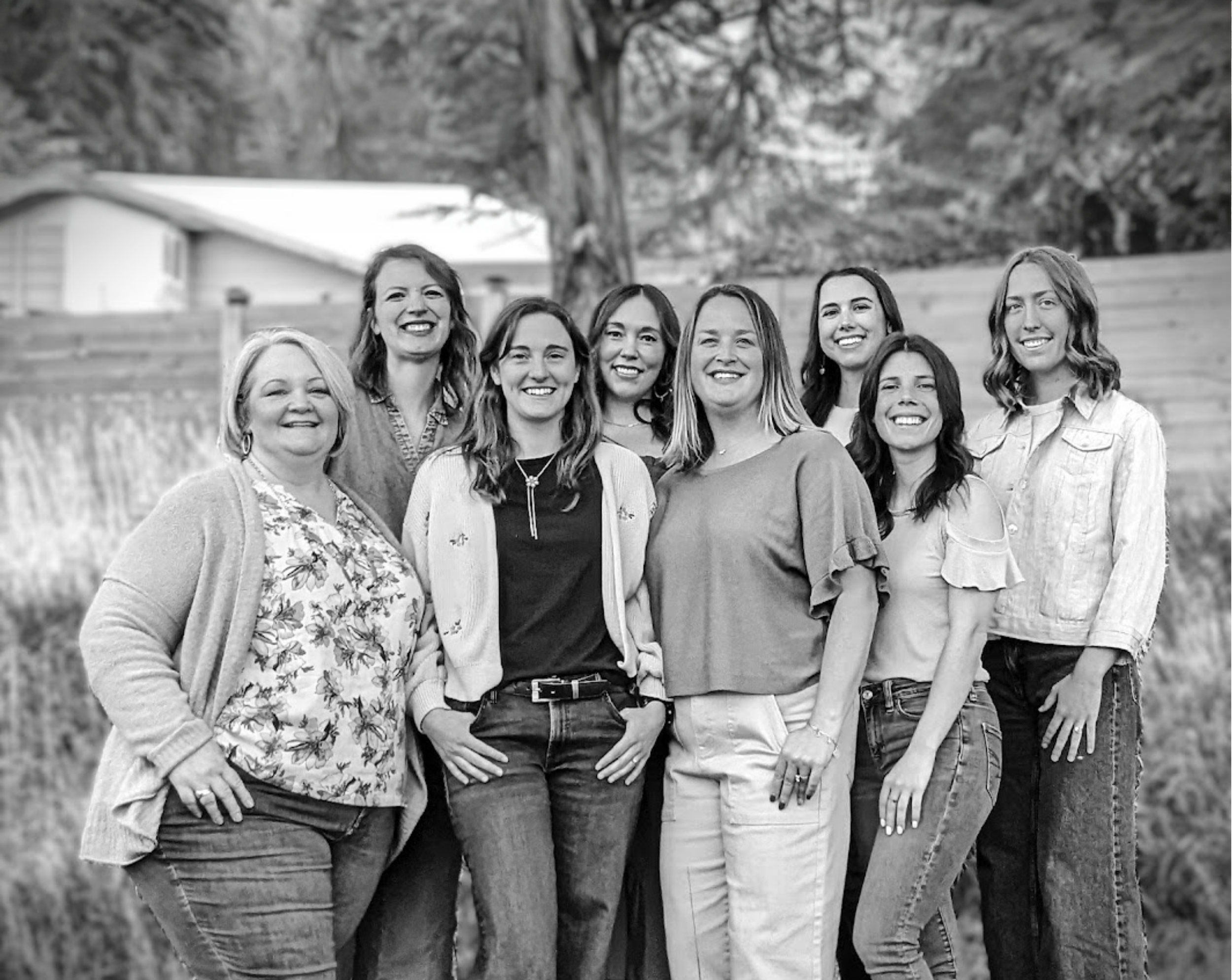 A group of nine women standing outdoors, smiling at the camera, with trees and a house in the background.