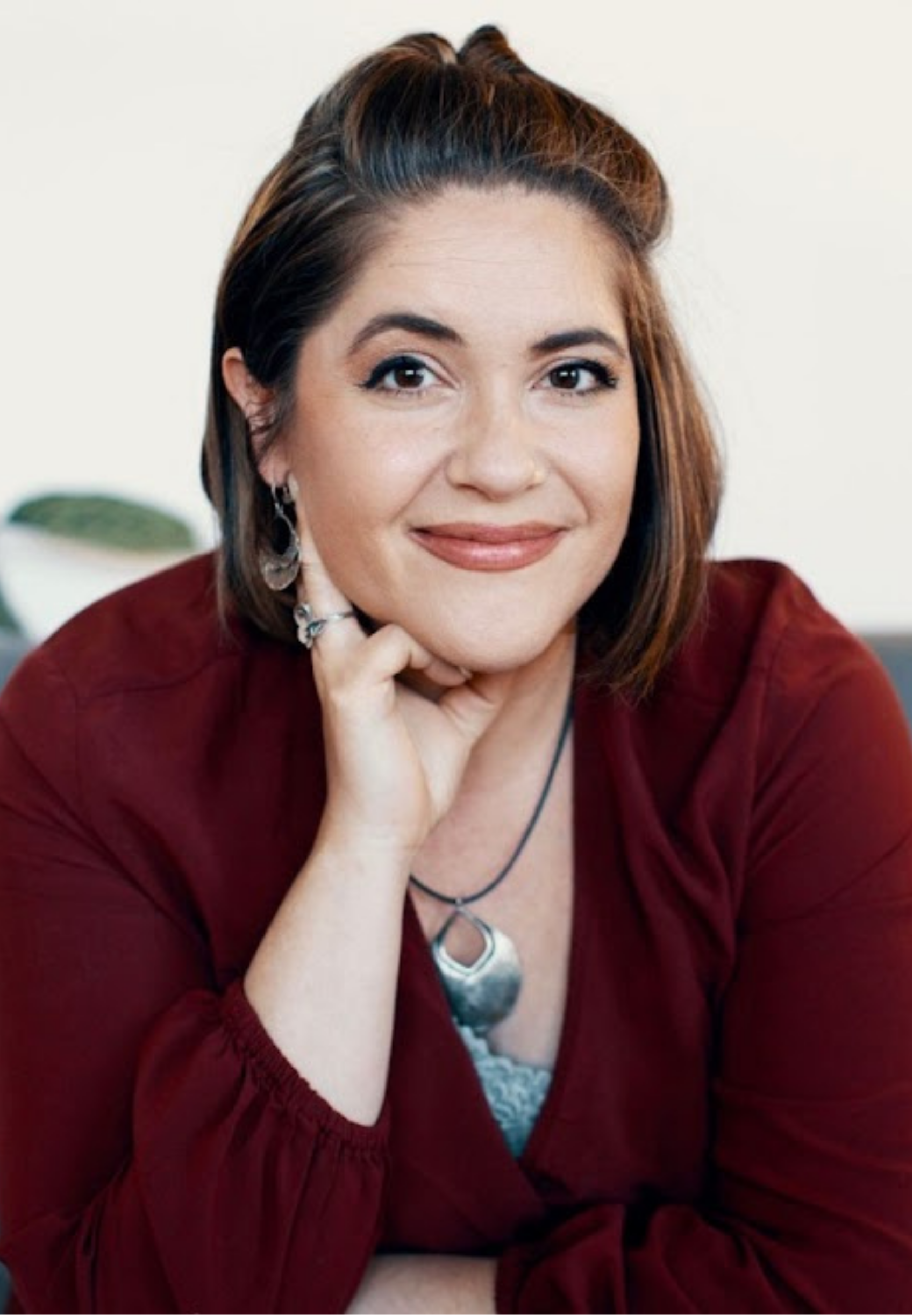 Angi Baker, with shoulder-length brown hair, wearing a red blouse, resting her chin on her hand with a smile, in a well-lit indoor setting.