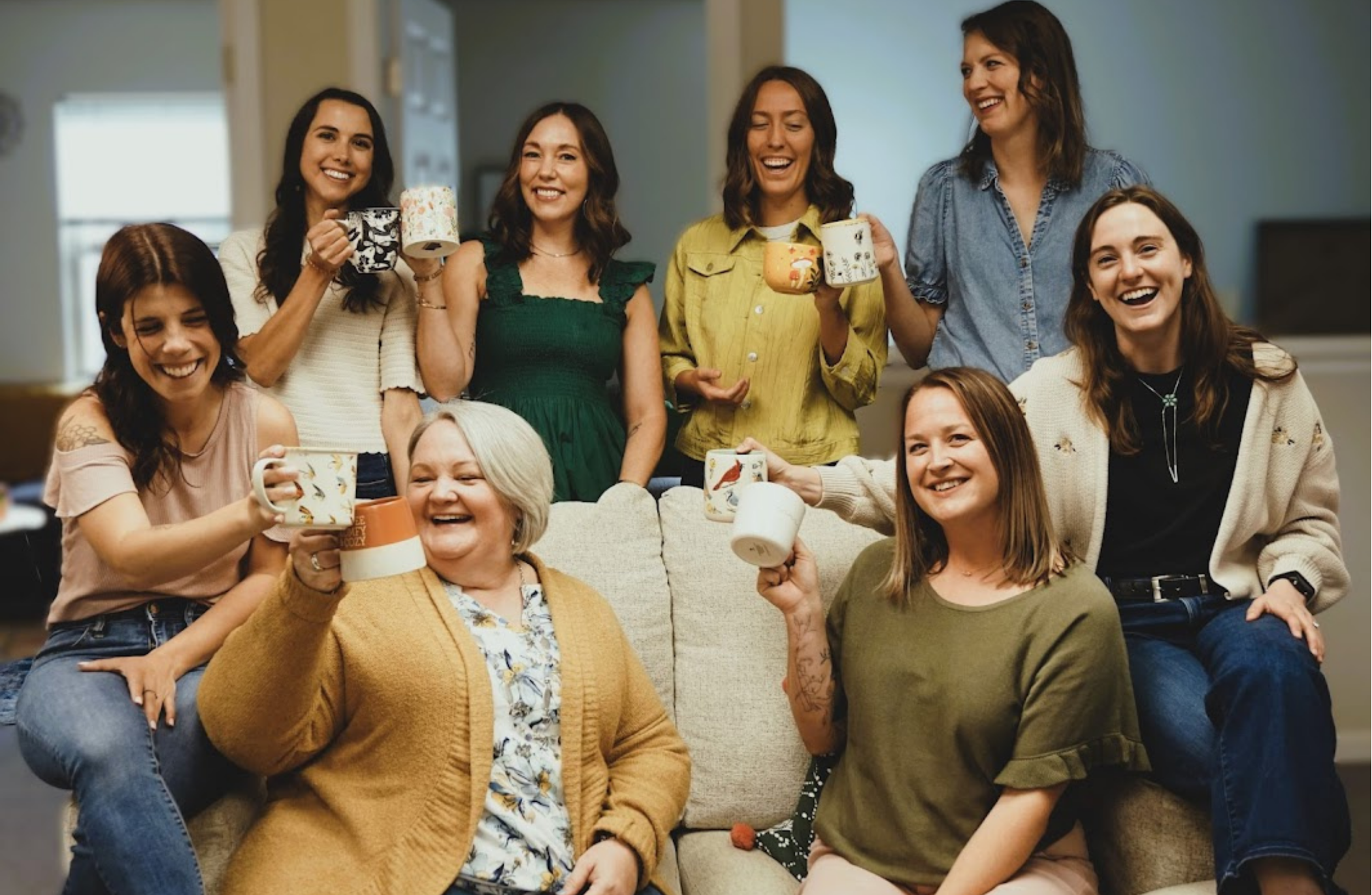 A group of women gathered together in a cozy living room, smiling and holding mugs of coffee or tea, celebrating and enjoying each other's company.