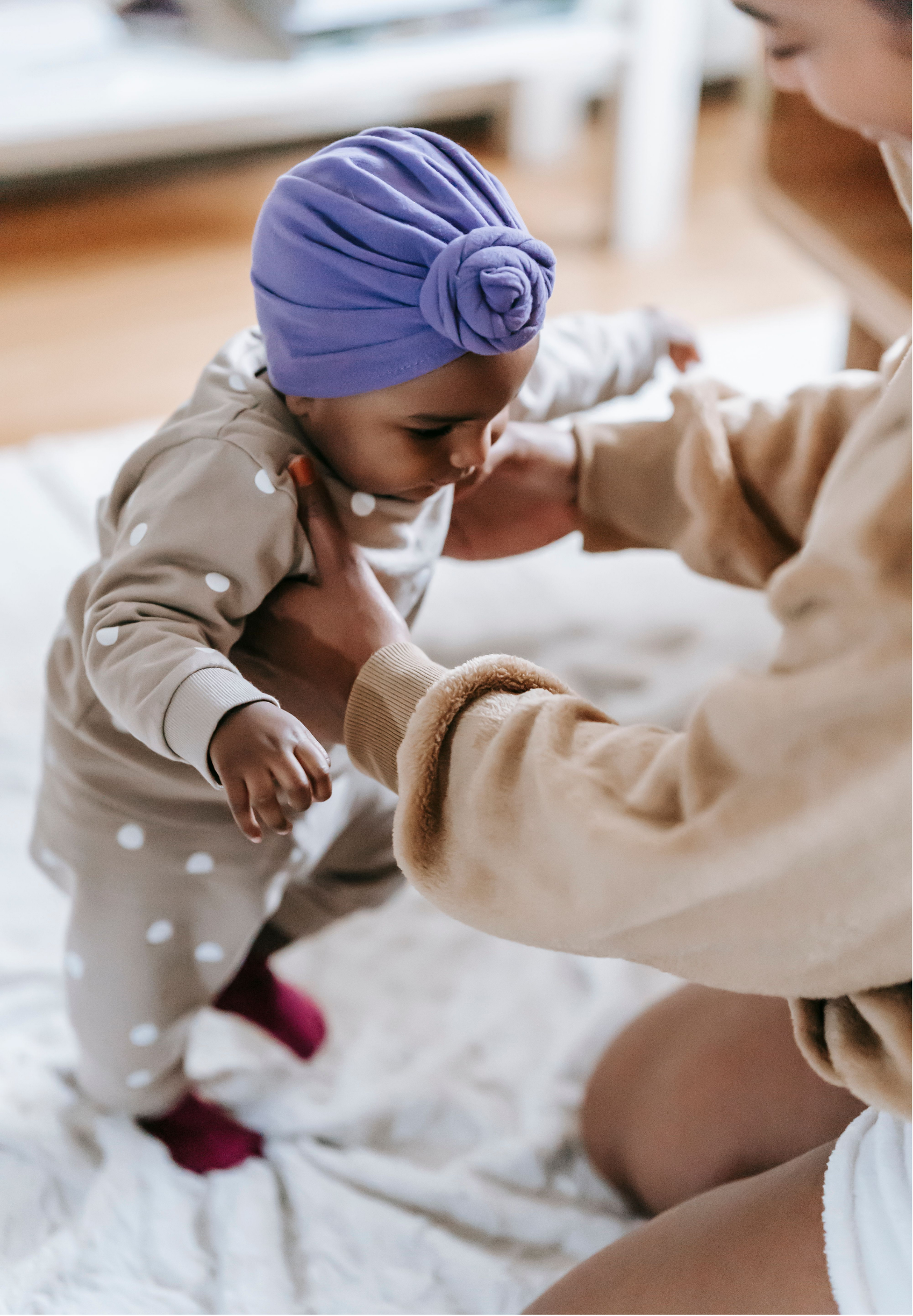 A young girl wearing a purple headscarf and polka dot pajamas is leaning forward with the help of an adult. They are on a white blanket or bedspread, and her face is close to someone's knee while her arms are outstretched. The scene appears to be indoors in a cozy setting.