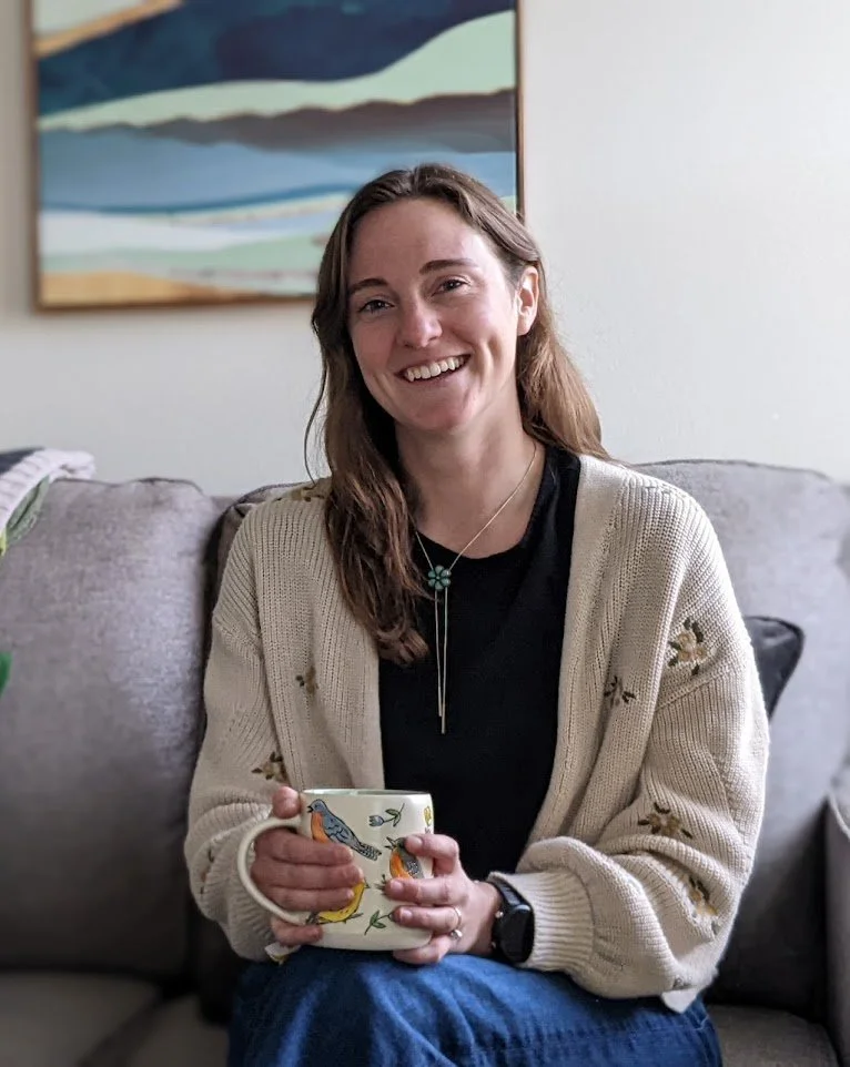 Melissa Ceren, therapist in Colorado Springs, sitting on a gray couch, holding a colorful mug, smiling, with a painting of a landscape hanging on the wall behind her.