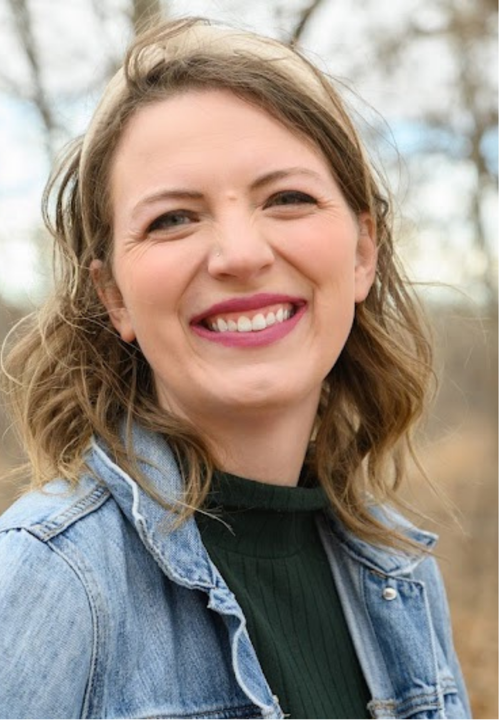 Jennie Radspinner, smiling outdoors, wearing a denim jacket over a black top, with blurred trees in the background.