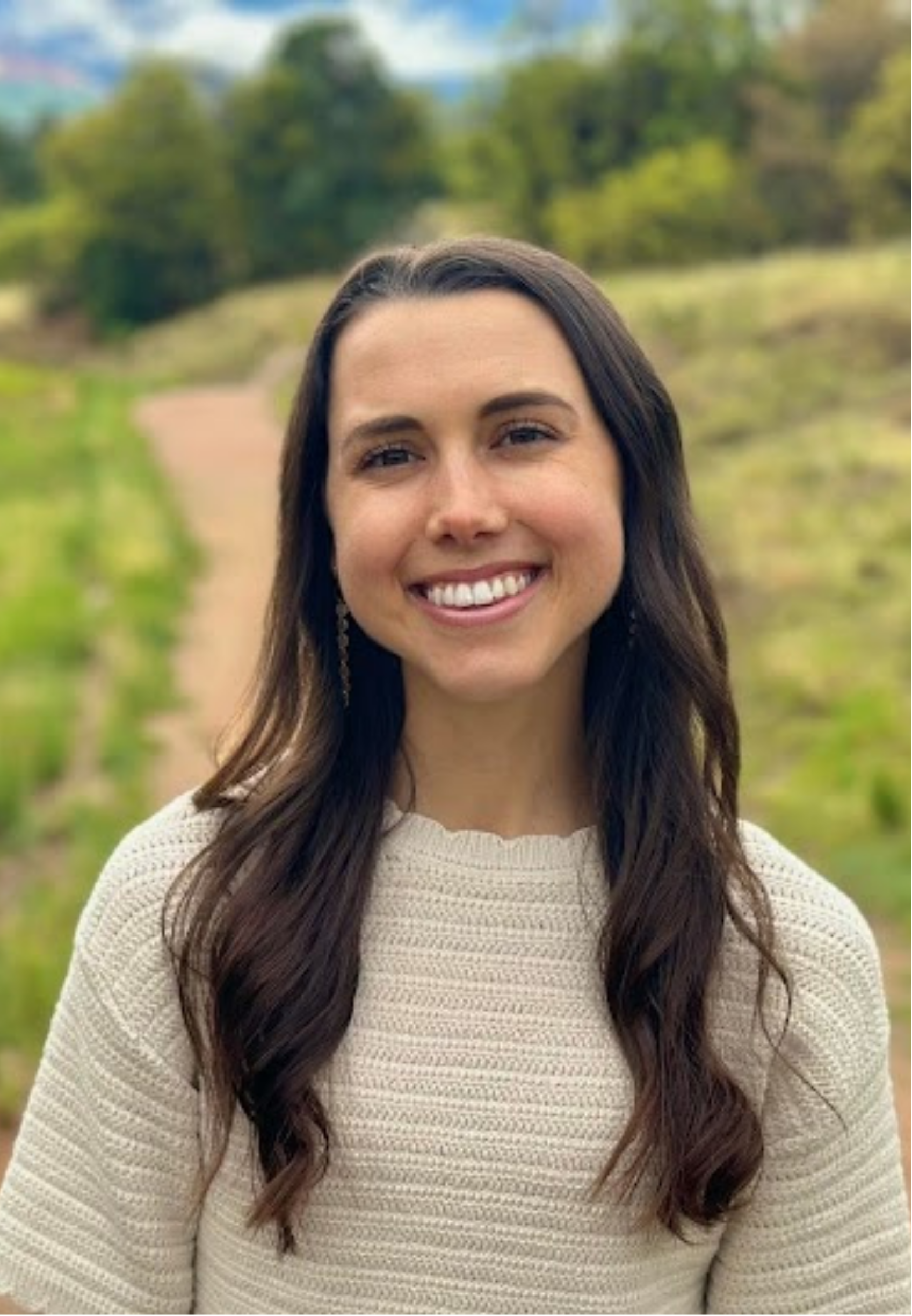 Alexa Beck, with long brown hair and a bright smile standing outdoors in a natural setting with trees and a trail in the background.