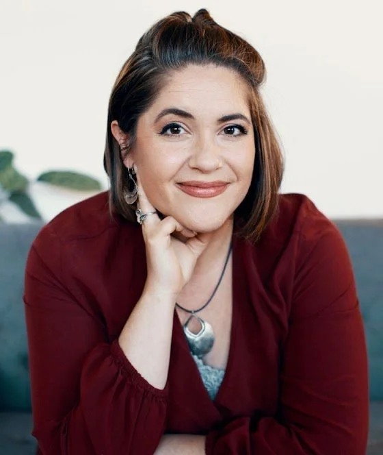 Angi Baker, with shoulder-length brown hair, wearing a burgundy top and earrings, smiling while resting her chin on her hand in an indoor setting.