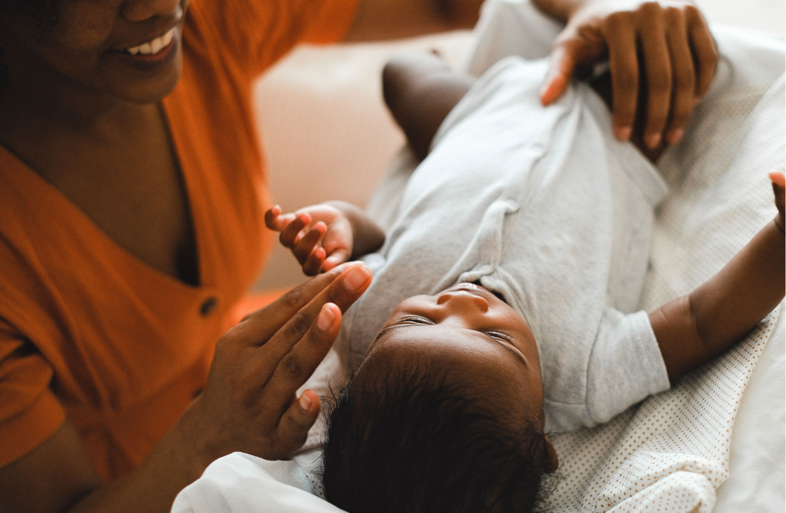 A person in an orange shirt smiling and gently touching a young child's face, who is lying down on a bed with eyes closed.