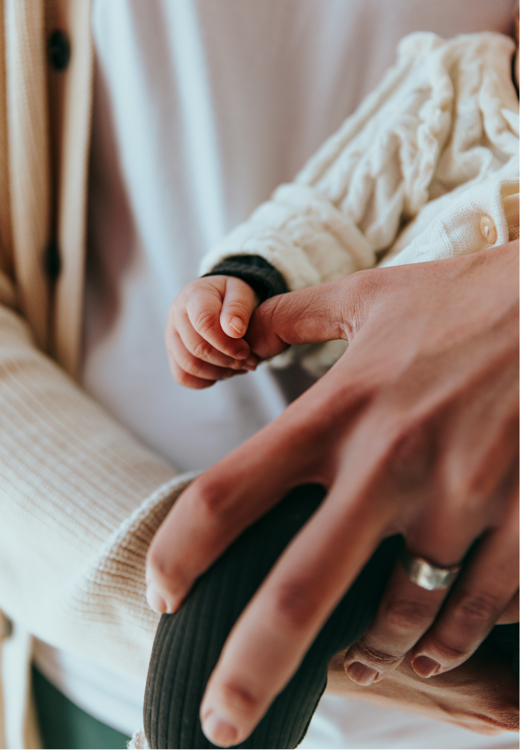 Close-up of a baby's hand holding an adult's finger, with the adult wearing a ring and a motorcycle-style glove. The baby's sleeve is a cream-colored cable knit sweater.