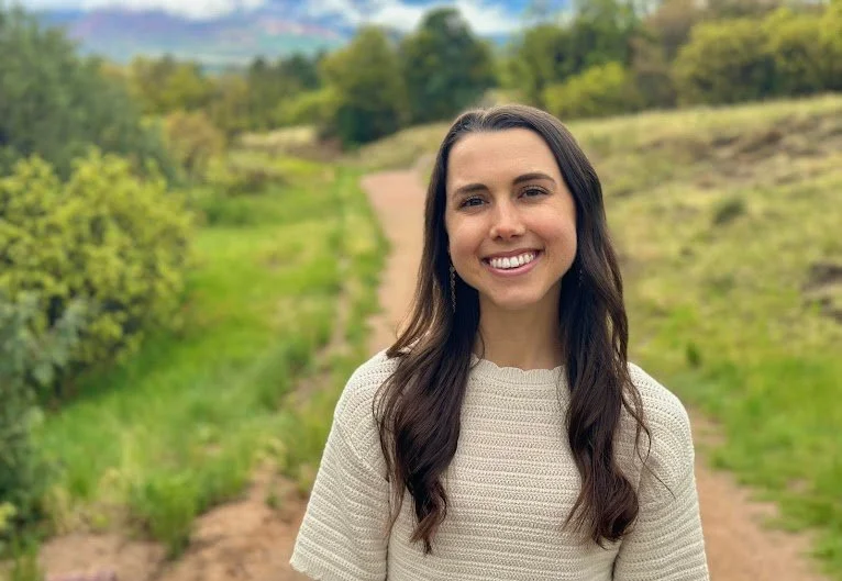 Alexa Beck, with long dark hair smiling outdoors on a dirt path surrounded by green trees and grass, with hills and a cloudy sky in the background.