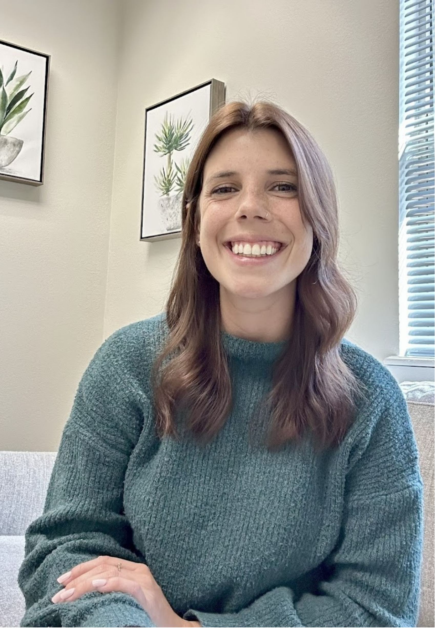 Colleen Morrison, smiling, with light brown wavy hair, wearing a teal sweater, sitting indoors near a window with blinds. There are framed botanical prints on the wall behind her.