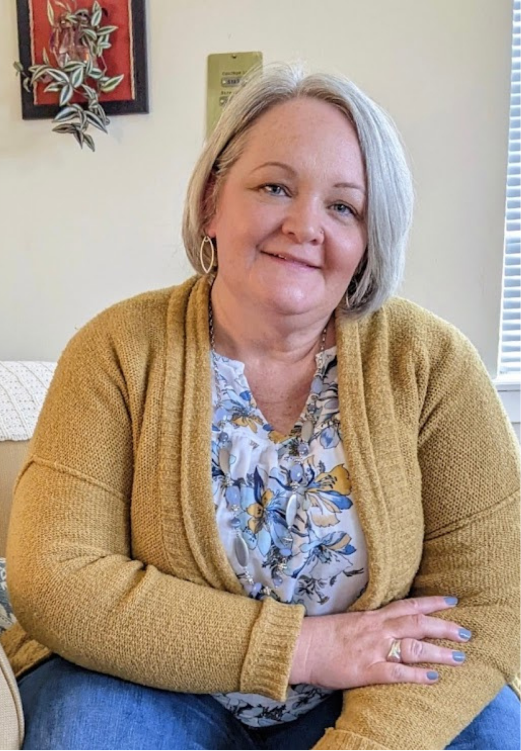Lou Ann Marquez, therapist in Colorado Springs, with short gray hair and earrings, wearing a mustard yellow cardigan and floral blouse, sitting with crossed arms.