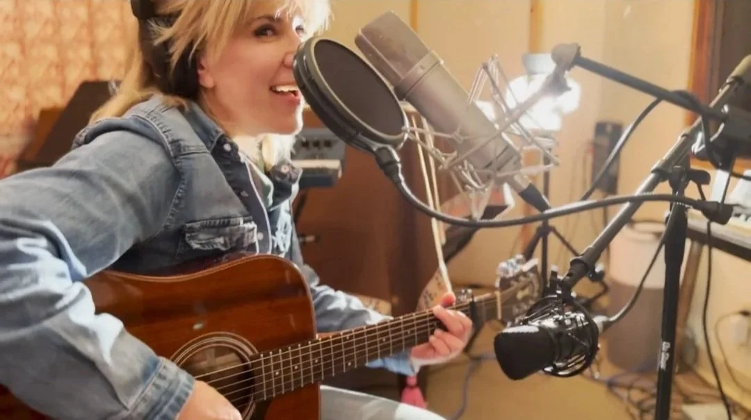 Amanda Pearcy playing an acoustic guitar in a music studio with a microphone in front of her.