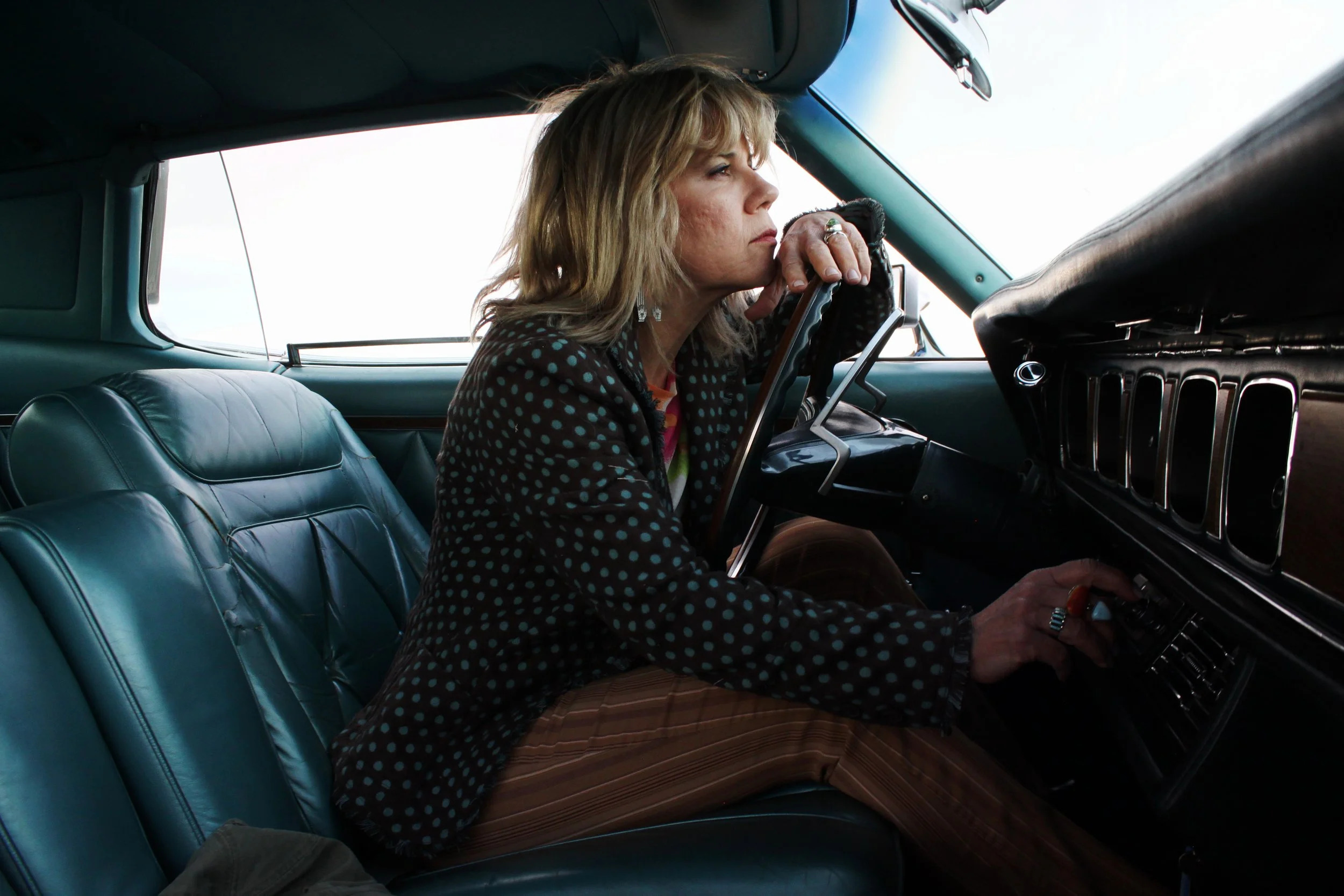 A woman with blonde hair sits inside a vintage car, resting her chin on her hand and looking ahead through the windshield, with a contemplative expression.