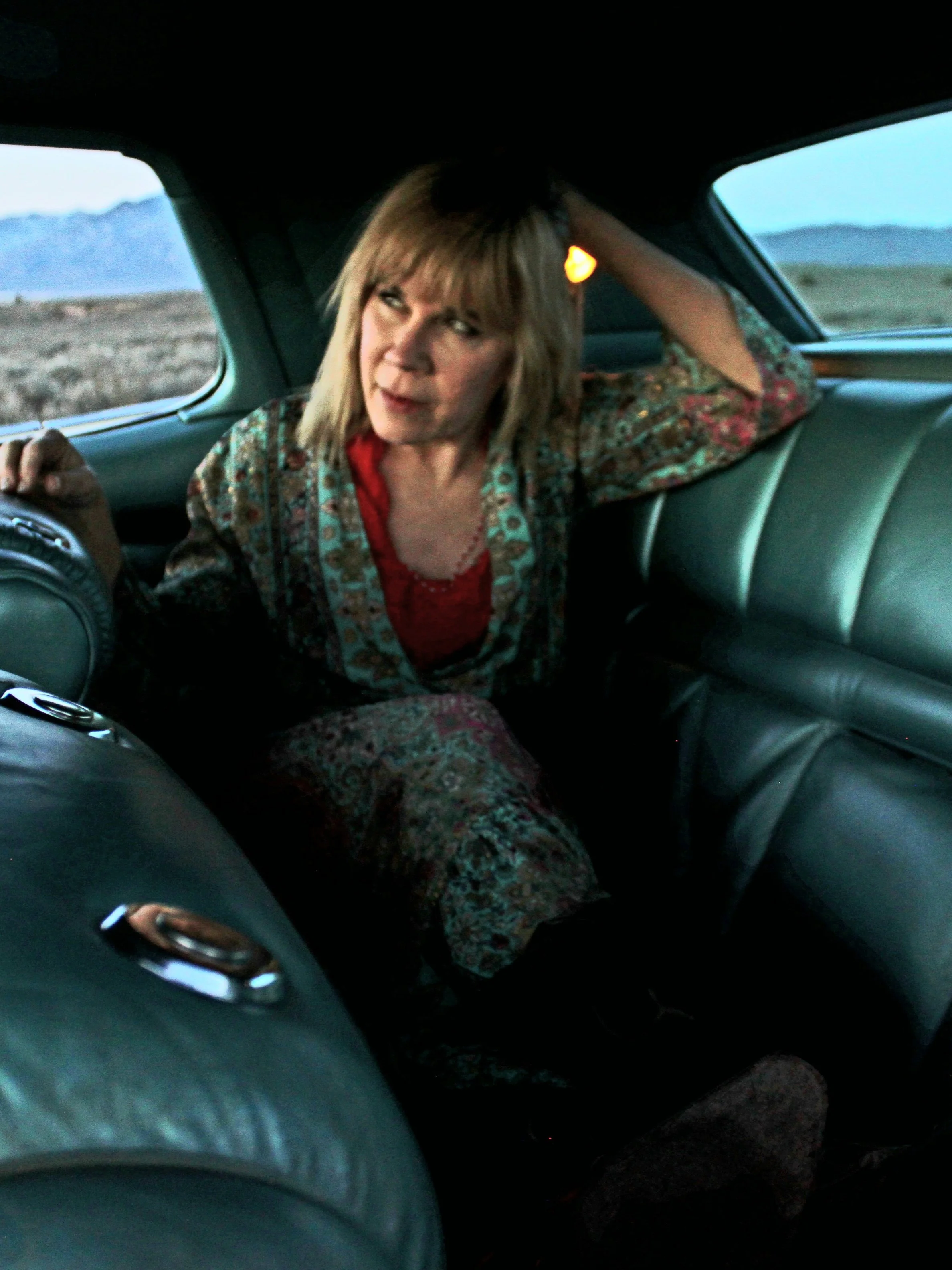 A woman with blonde hair and bangs sitting in the backseat of a car, leaning her head on her hand, looking at the camera, wearing a patterned jacket over a red top, with mountains visible through the windows.