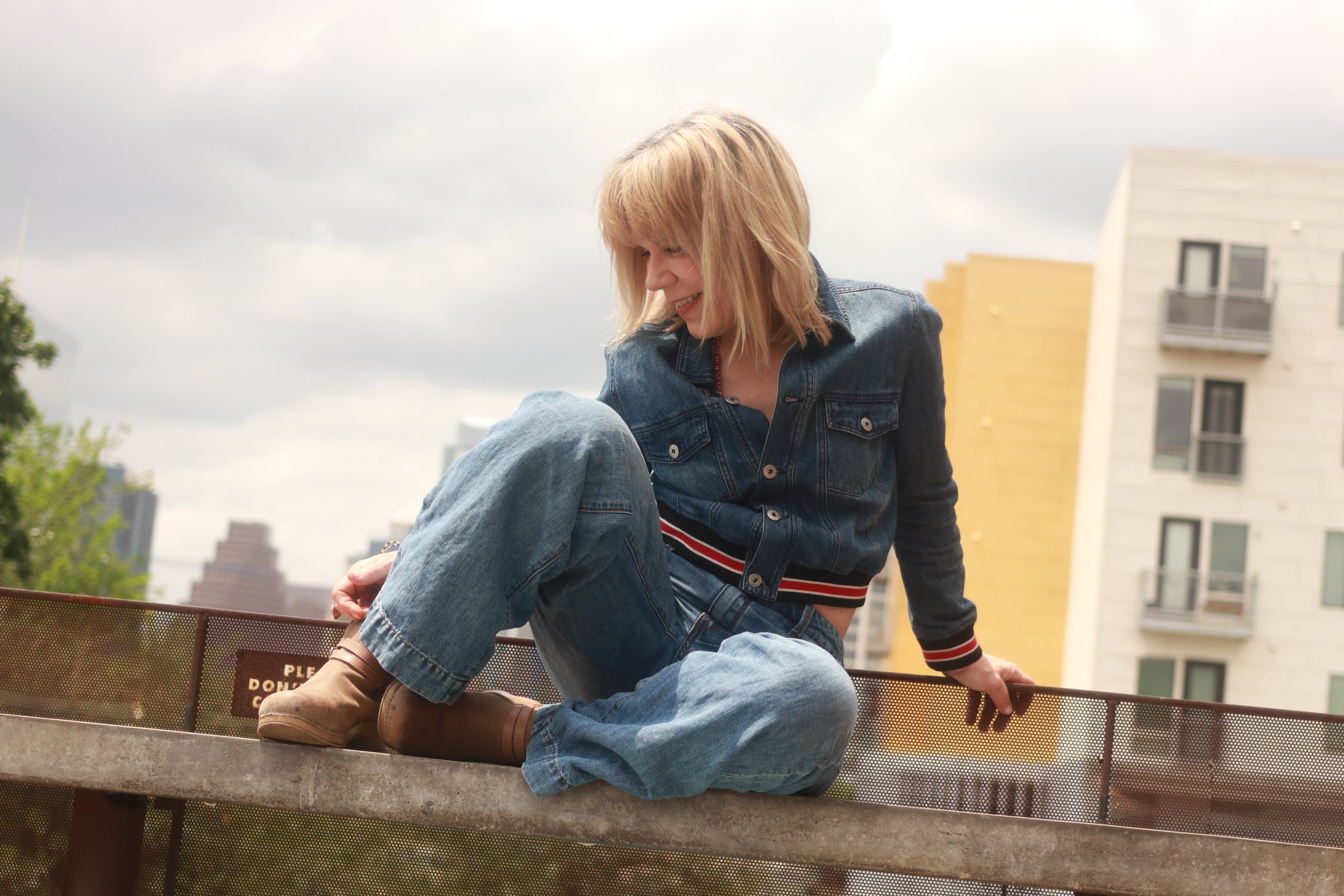 Amanda Pearcy smiling woman with blond hair, wearing a denim jacket and striped shirt, sitting on a metal railing outdoors with city buildings in the background under a cloudy sky.