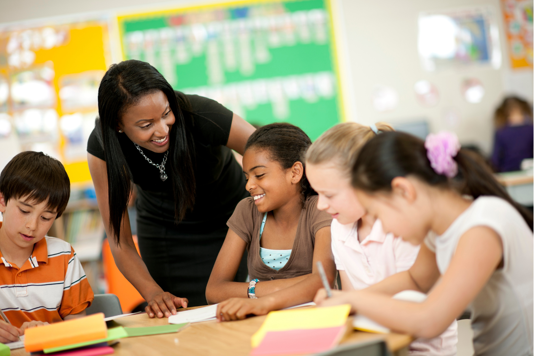 A teacher coaching students in a classroom with colorful bulletin boards in the background. The students are focused on their work at a table.