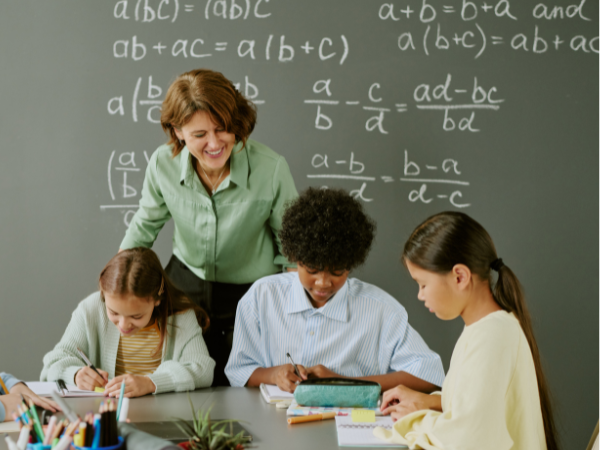 A teacher smiling and leaning over a table with three young students, working on homework or assignments in a classroom with a blackboard filled with mathematical equations in the background.
