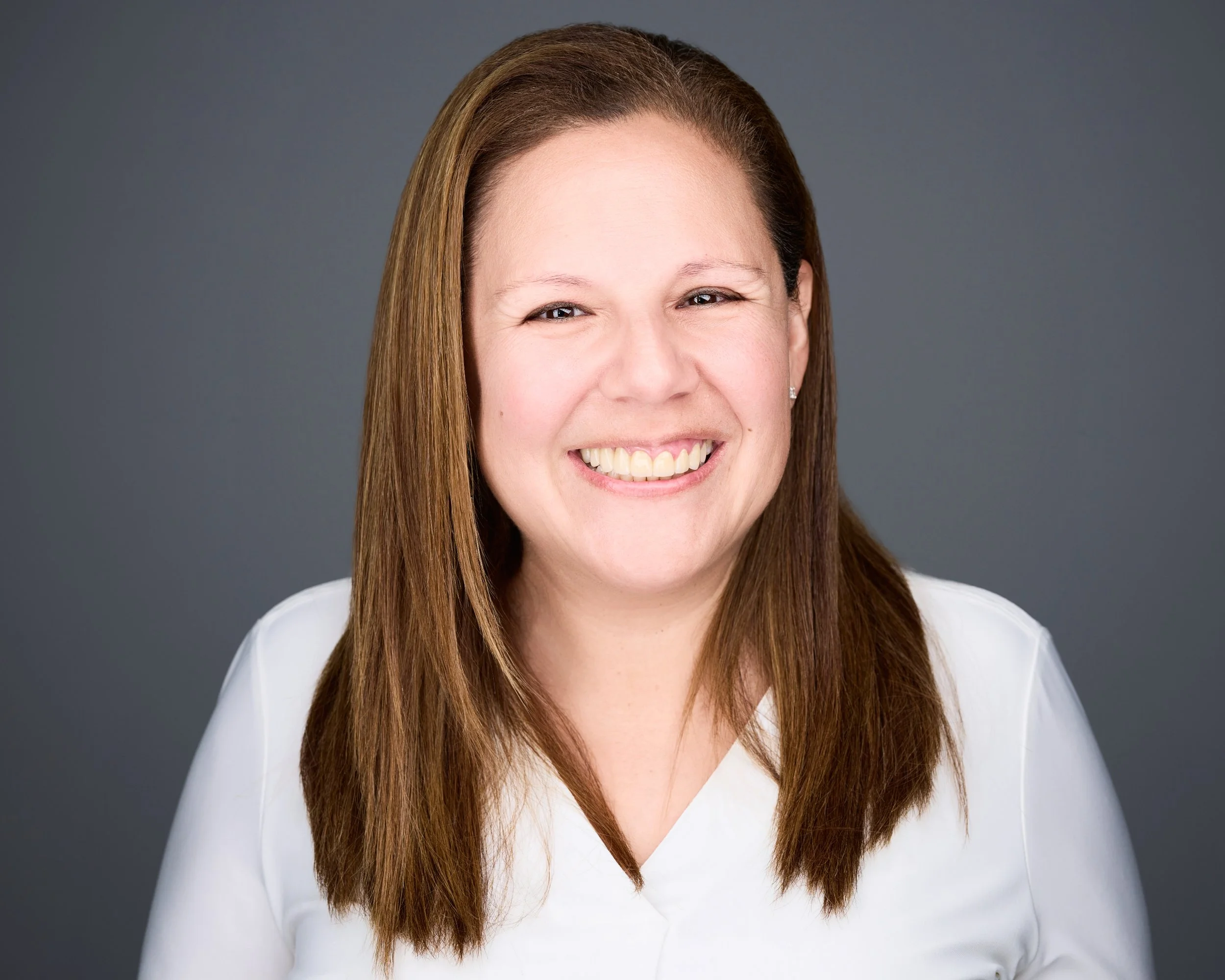 Close-up portrait of a smiling woman with long brown hair wearing a white top against a gray background.