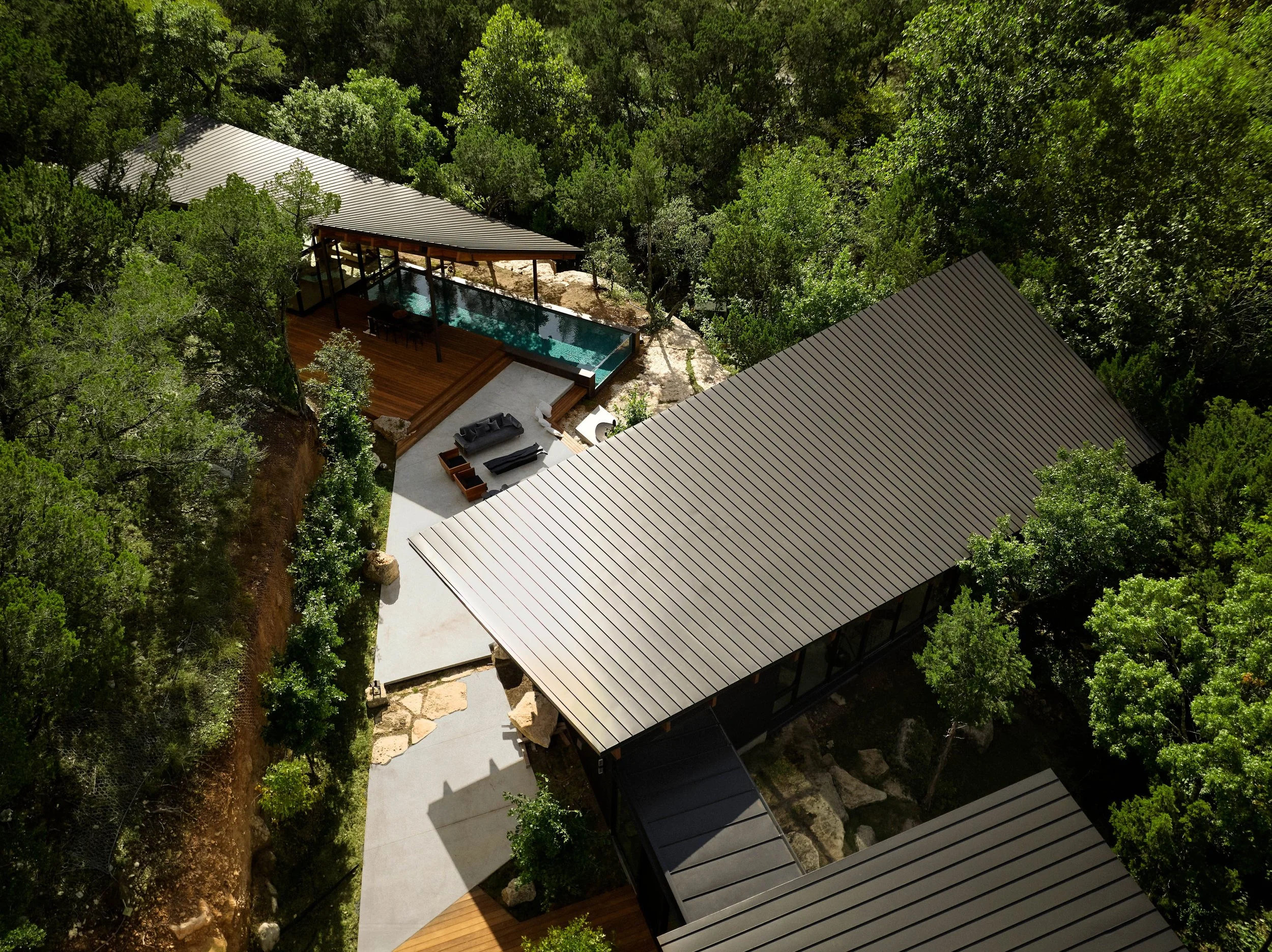 Aerial view of a modern house with a metal roof, surrounded by dense green trees, with a wooden deck, a small pool, and outdoor furniture.