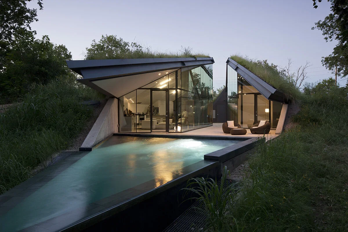 Modern house with glass walls and sloped grass-covered roofs, situated on a hillside with greenery, featuring an outdoor swimming pool in the front with mood lighting and outdoor seating area.