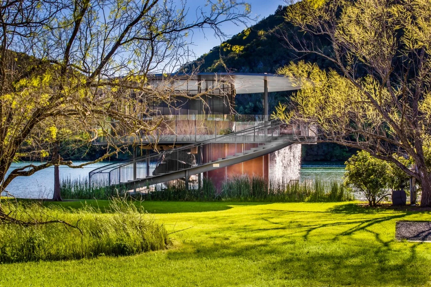 Modern building with glass walls on stilts over water, surrounded by trees and a grassy lawn with shadows from the trees, in a natural setting with hills and water in the background.