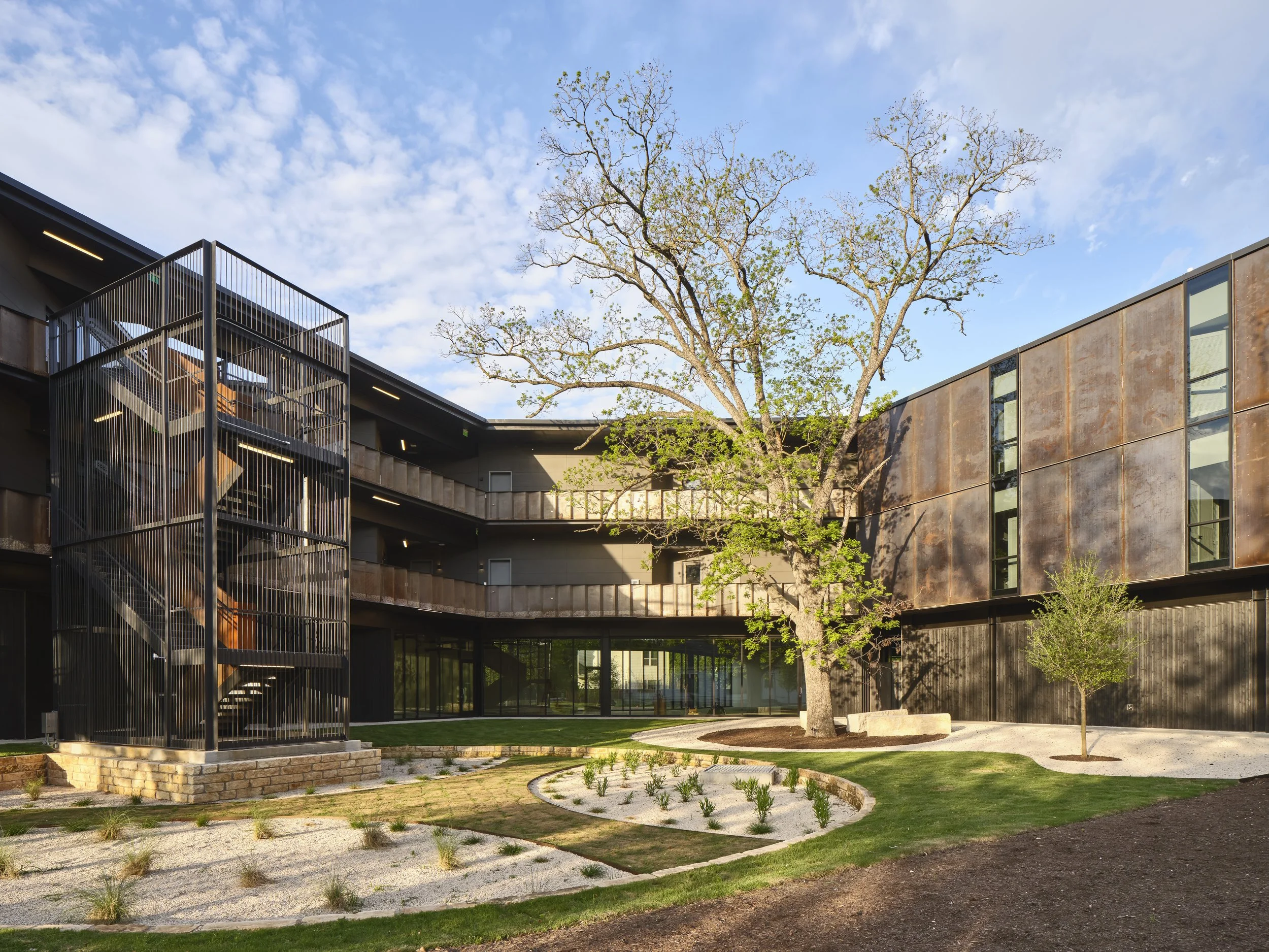 Modern building with outdoor staircase, surrounded by a landscaped yard with a large tree with budding leaves and a blue sky with clouds.