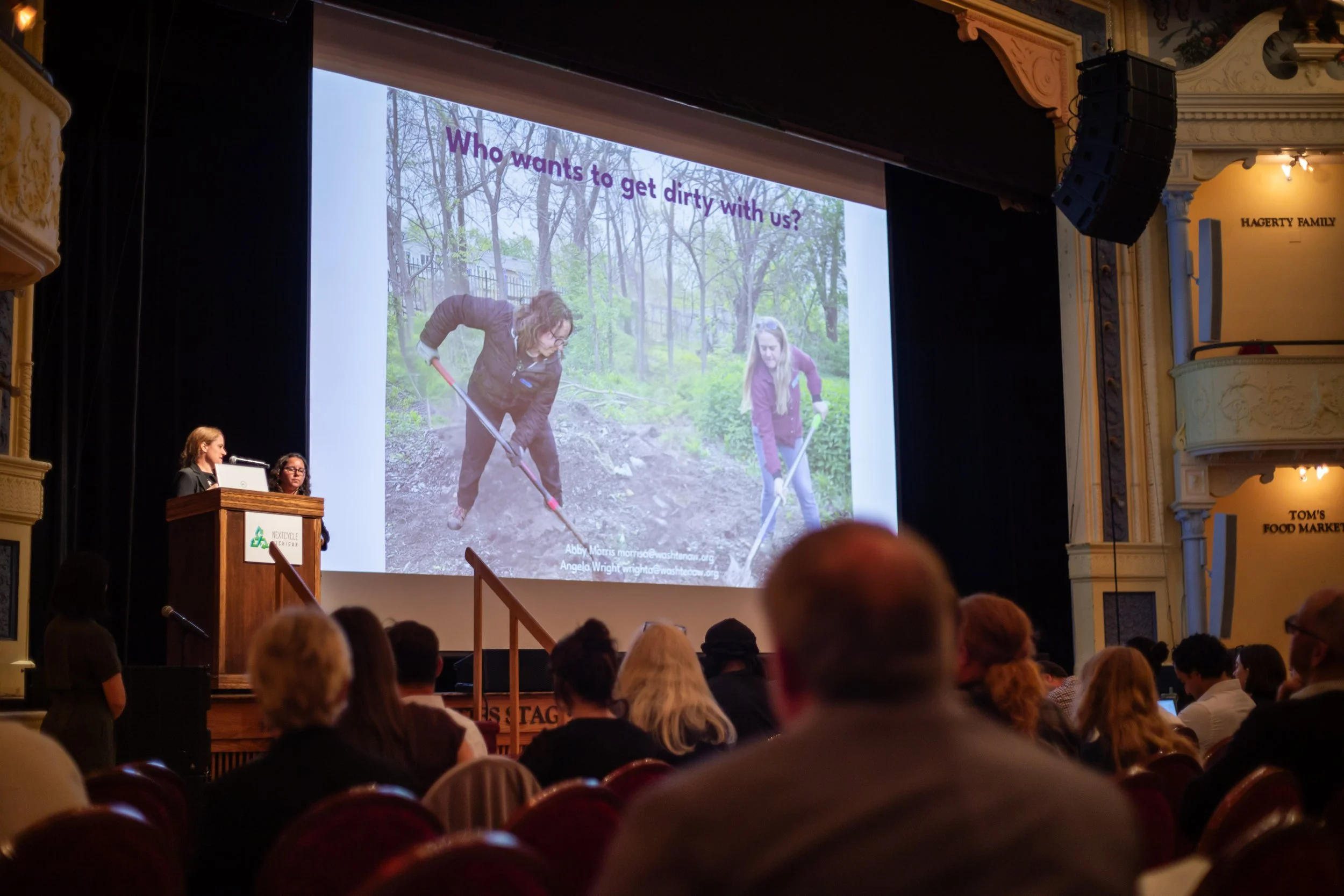 Two women giving a presentation to a crowd with a photo of people shoveling in the background.