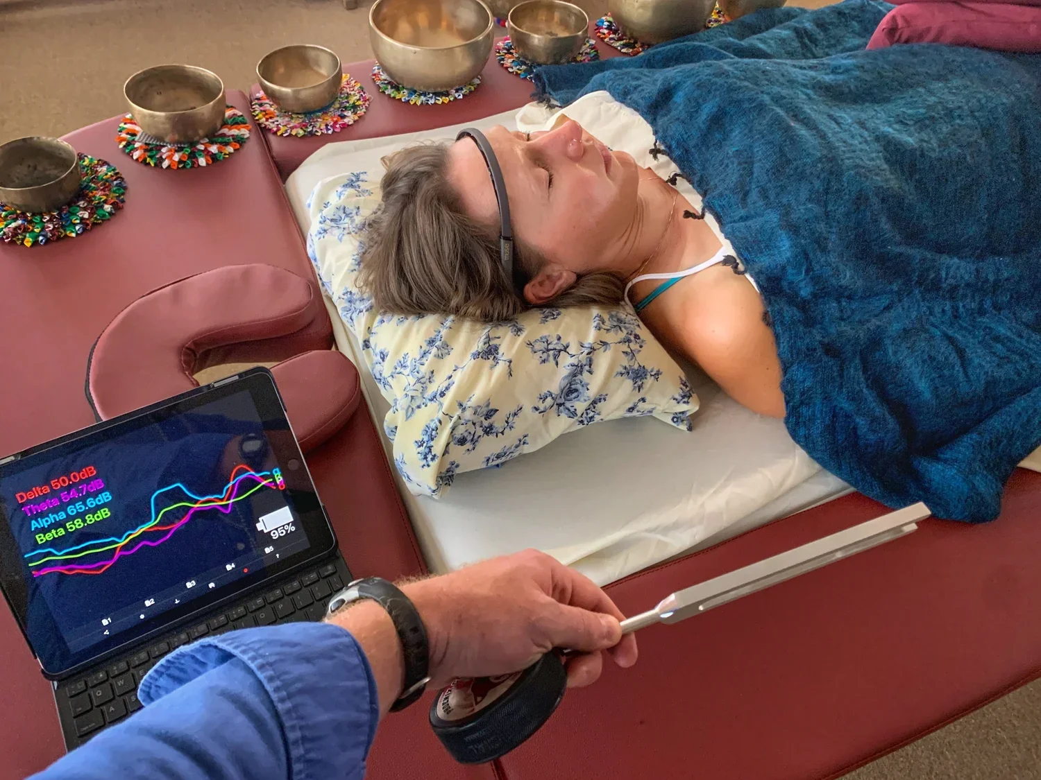 A woman with glasses lying on a bed with her eyes closed, covered with a blue blanket, and resting her head on a floral pillow. A person in a blue shirt is holding a stethoscope to her chest while a laptop displaying brain wave activity is nearby, showing colored lines and data.