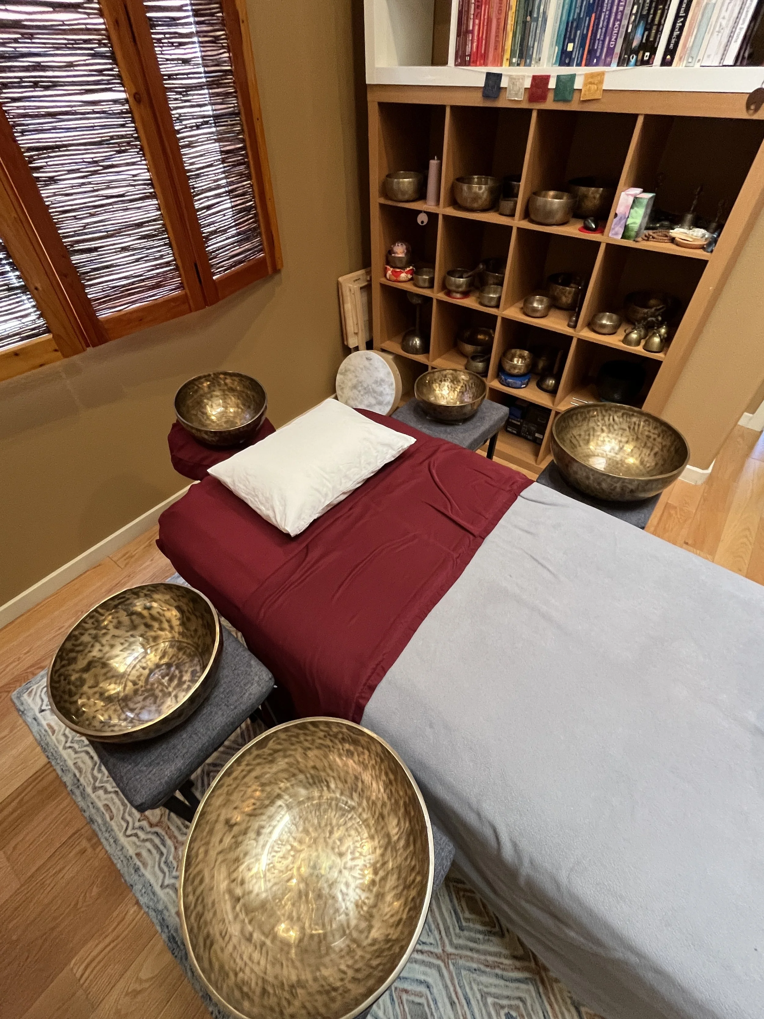A room with a massage table covered with a gray sheet and a red blanket, surrounded by 10 brass singing bowls of various sizes, some on small stools and others on the floor. There is a shelf with more singing bowls and books, and a window with wooden blinds.