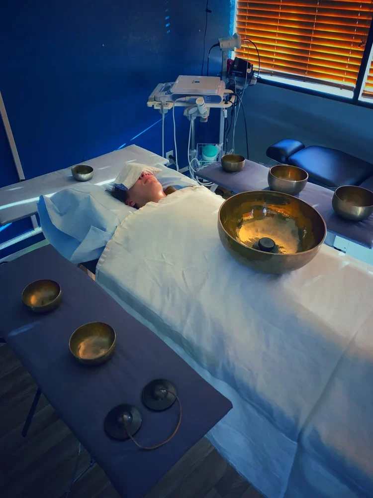 A person lying in a hospital bed with a towel over their forehead, surrounded by singing bowls, in a room with blue walls, wooden blinds, and medical equipment.