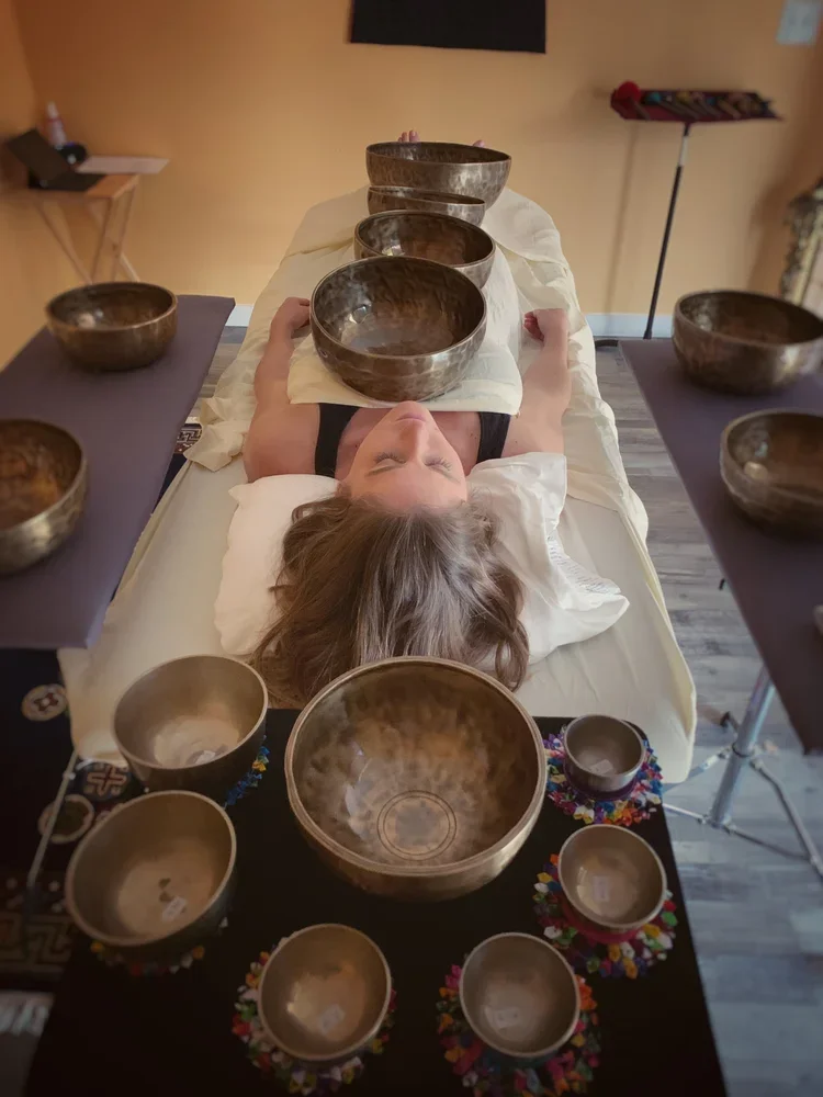 A woman lying on a massage table with bronze singing bowls surrounding her, used for sound therapy or meditation, in a wellness or therapy room.