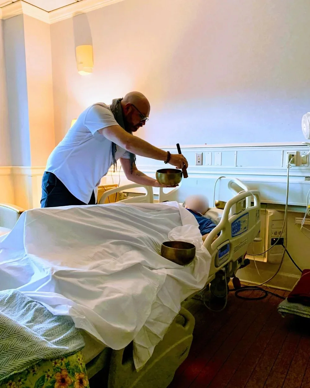 A man performing a spiritual ritual over a patient lying in a hospital bed, with bowls and a singing bowl on the bed, in a hospital room.