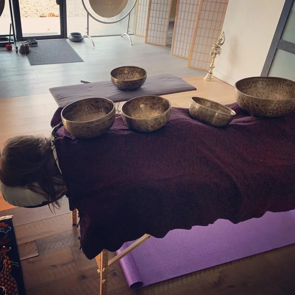 A massage or relaxation room with a massage table covered in a dark cloth, with large singing bowls and a purple yoga mat nearby, and an exercise area with weights, a meditation gong, and a partition in the background.