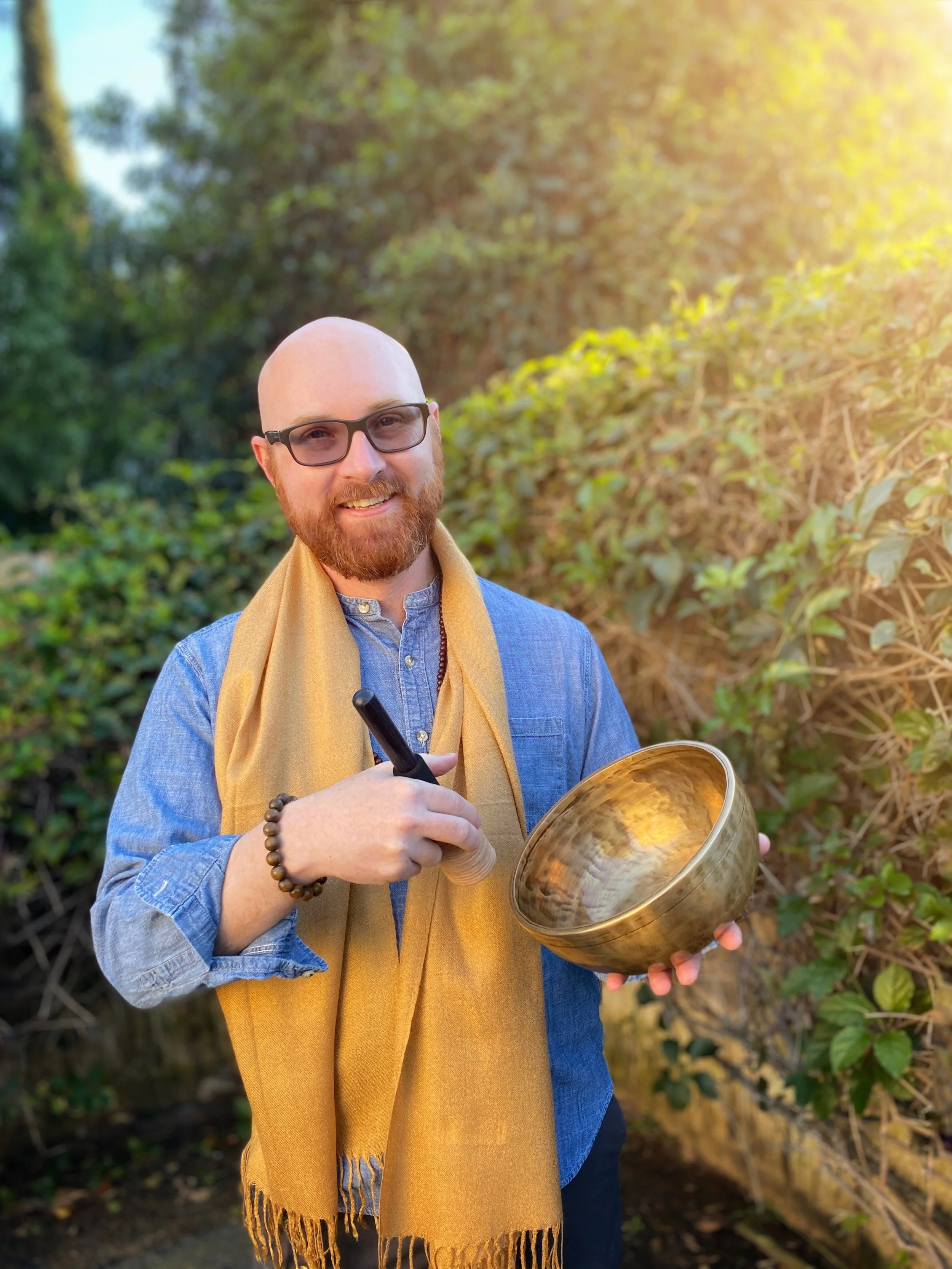 A man with glasses, a beard, and a bald head smiling while holding a Tibetan singing bowl and a mallet outdoors in a garden with green foliage and sunlight.