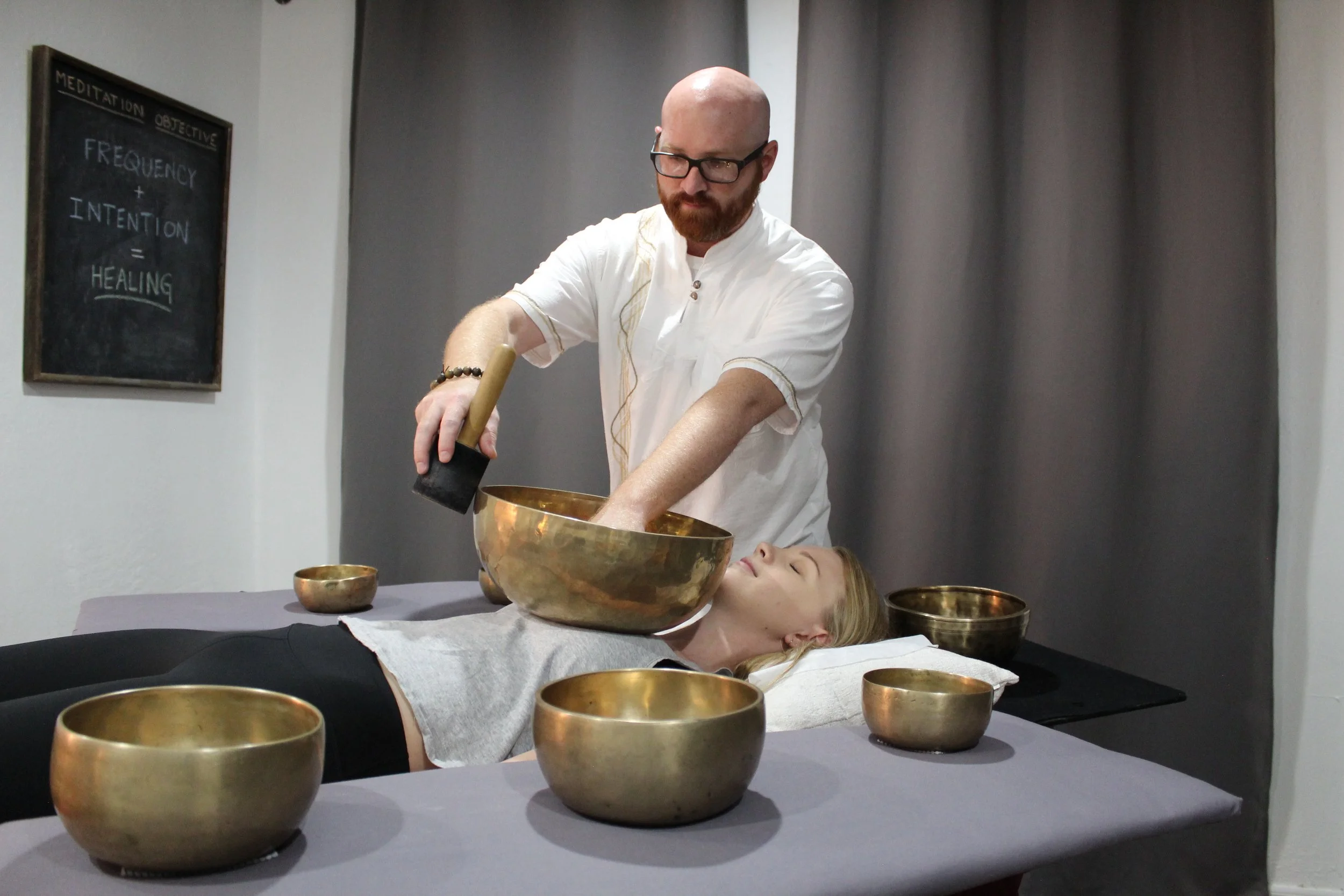 A person with long blonde hair is lying on a table, covered with a gray cloth. Several singing bowls are placed around her, and a man with glasses and a beard, wearing white, is holding a mallet and striking one of the bowls. A chalkboard on the wall has the words 'FREQUENCY + INTENTION = HEALING' written on it.