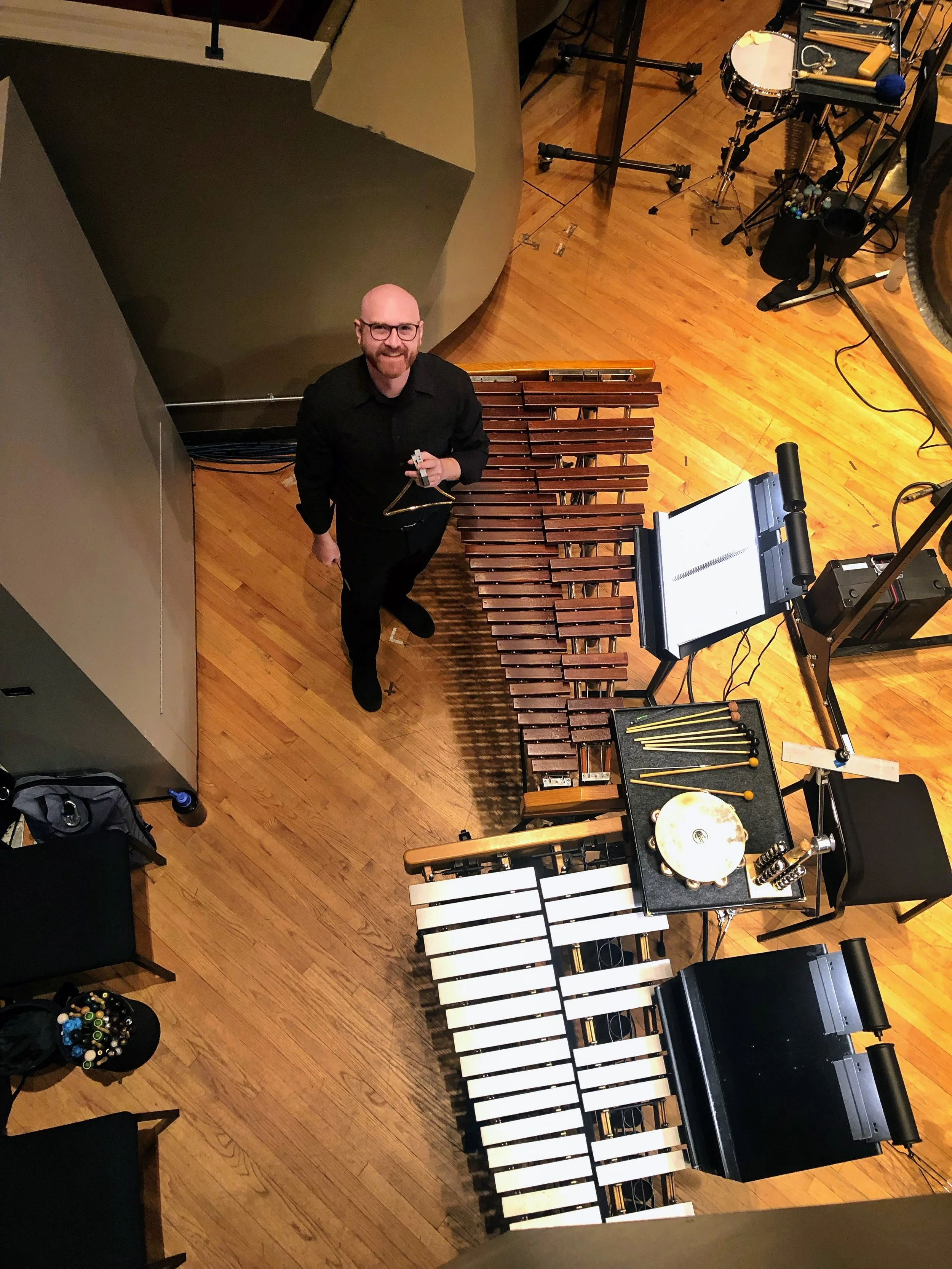 An overhead view of a man standing on a wooden floor, holding a triangle, smiling at the camera. Around him are musical instruments and equipment including a marimba, percussion instruments, a music stand with sheet music, and various percussion tools.