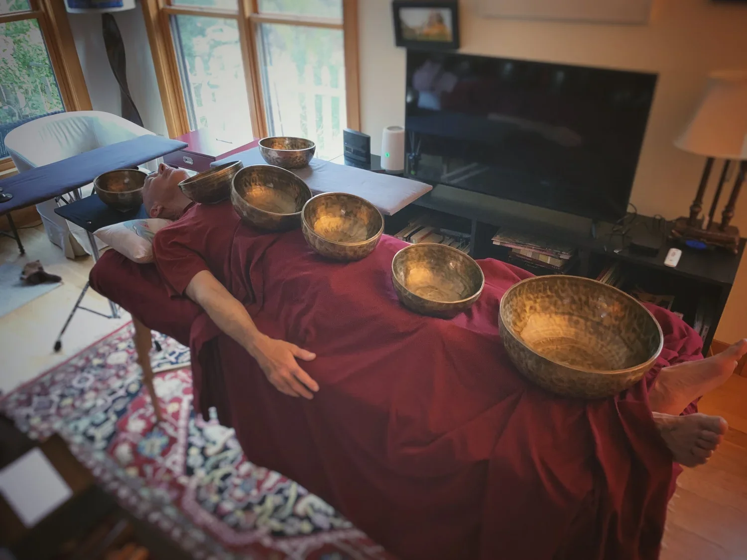 A person lying on a couch with multiple singing bowls placed on their body, in a cozy living room near large windows and a television.