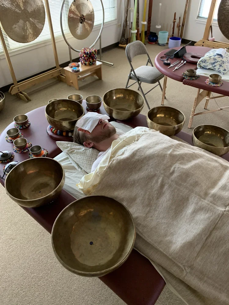 A person lying on a massage table with a cloth covering them and a blindfold over their eyes, surrounded by singing bowls of various sizes, in a room with healing or meditation objects.