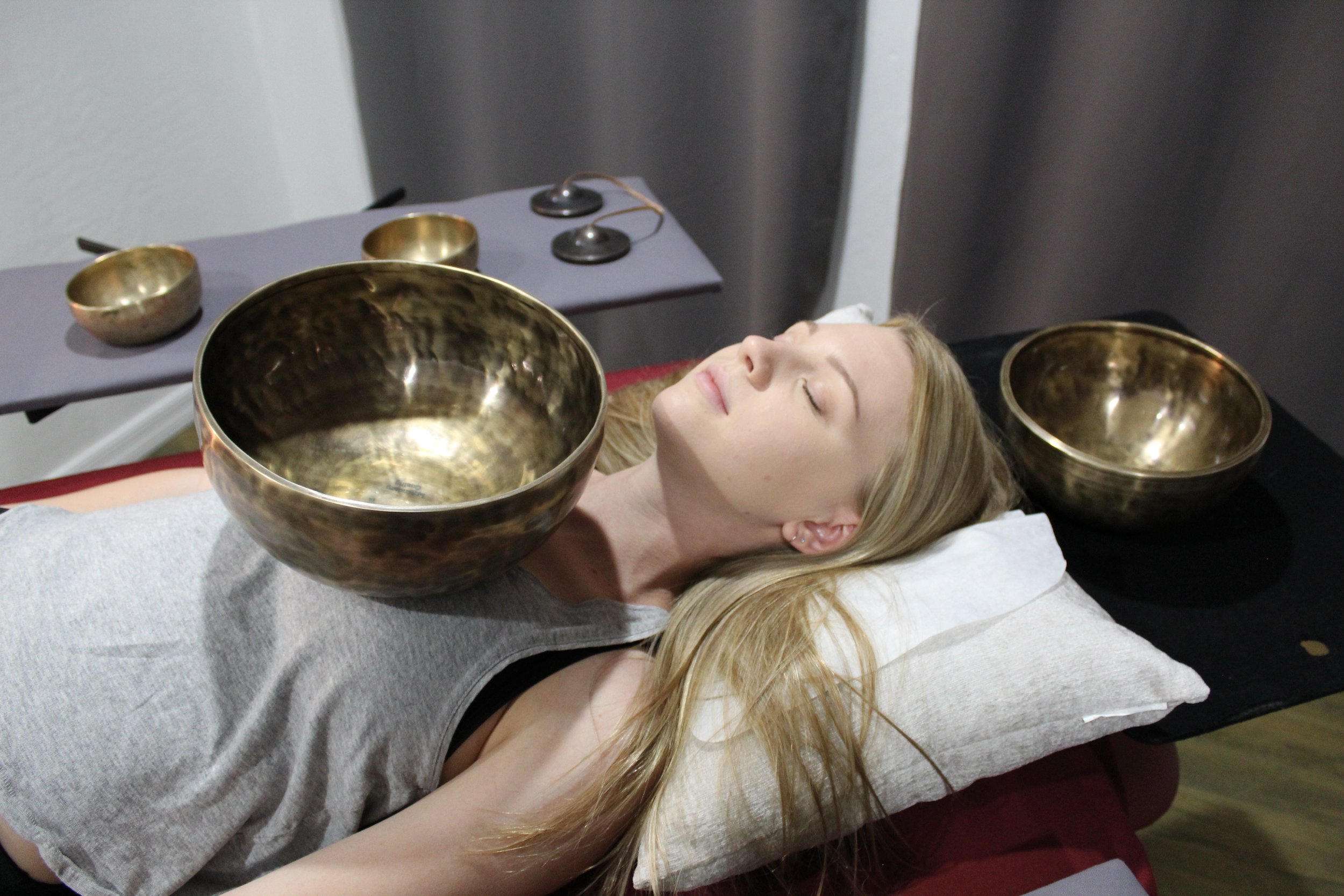 A woman lying on a massage table with her eyes closed, holding a singing bowl on her chest, with two smaller bowls and two singing bowls on a nearby table.