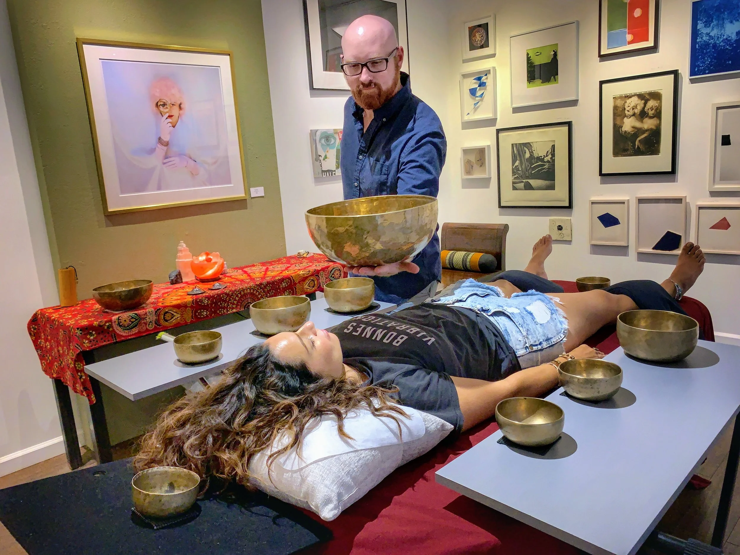 A man with glasses and a beard is holding a large brass singing bowl in an art gallery. He is standing behind a table with several smaller brass bowls and a red patterned cloth. Two women are lying down on tables with eyes closed, surrounded by bowls. The gallery wall behind features various framed artworks, including a large colorful portrait of a woman with pink hair.