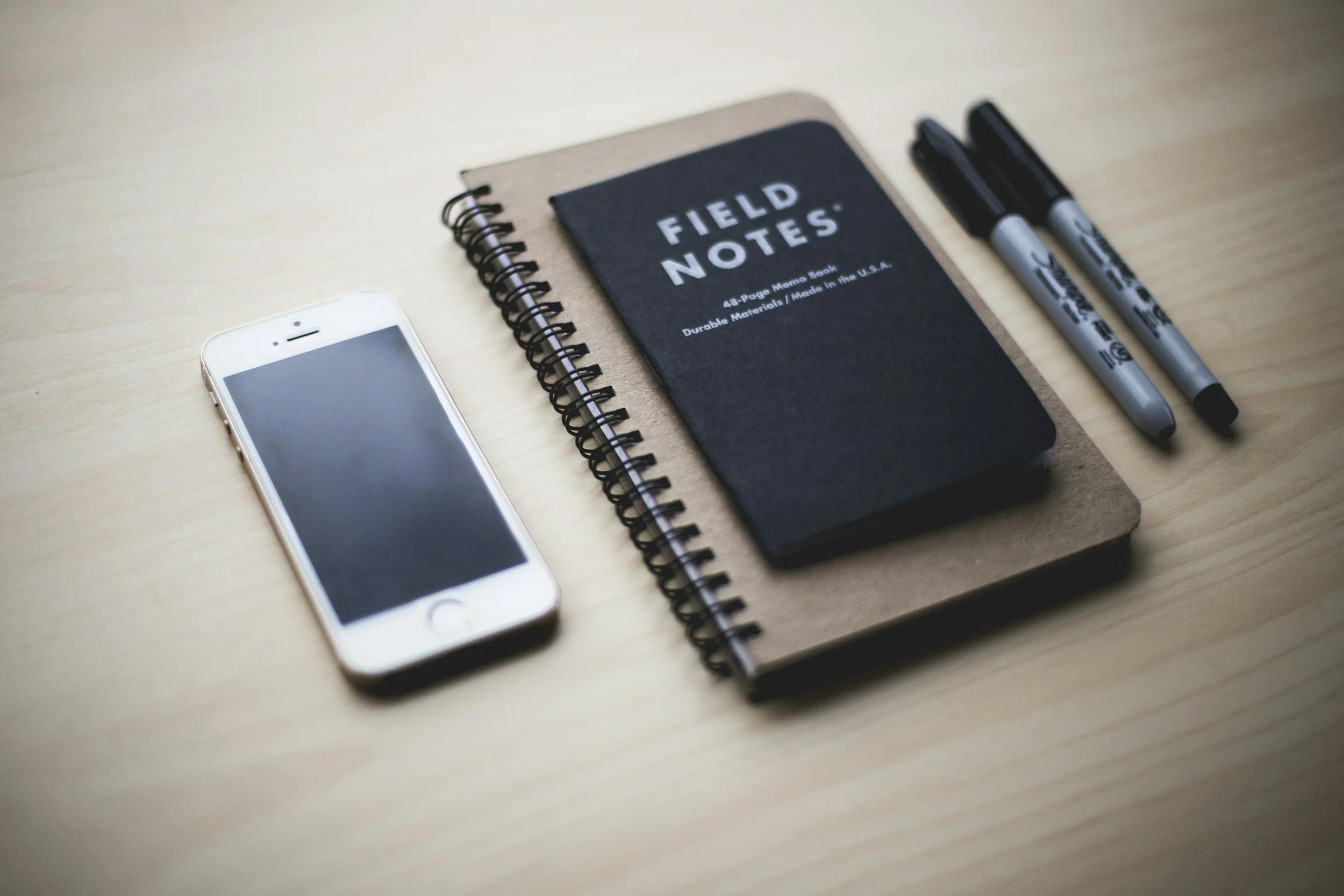 A wooden desk with a smartphone, a spiral-bound notebook labeled 'Field Notes', two black and gray Sharpie markers, and two black Pens.
