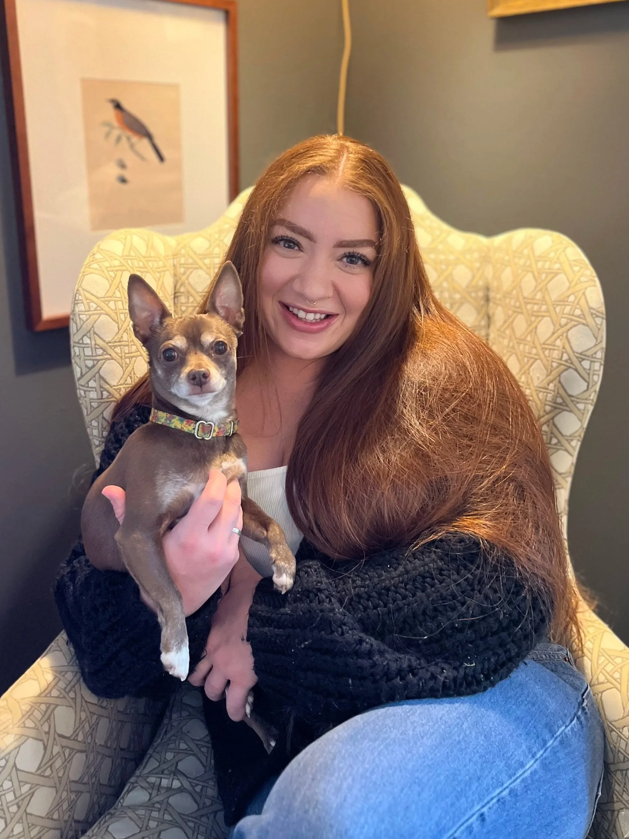 A woman with long red hair and a septum piercing smiling while holding a small brown Chihuahua dog with large ears on her lap. She is sitting in a patterned chair in a room with a framed picture of a bird on the wall behind her.