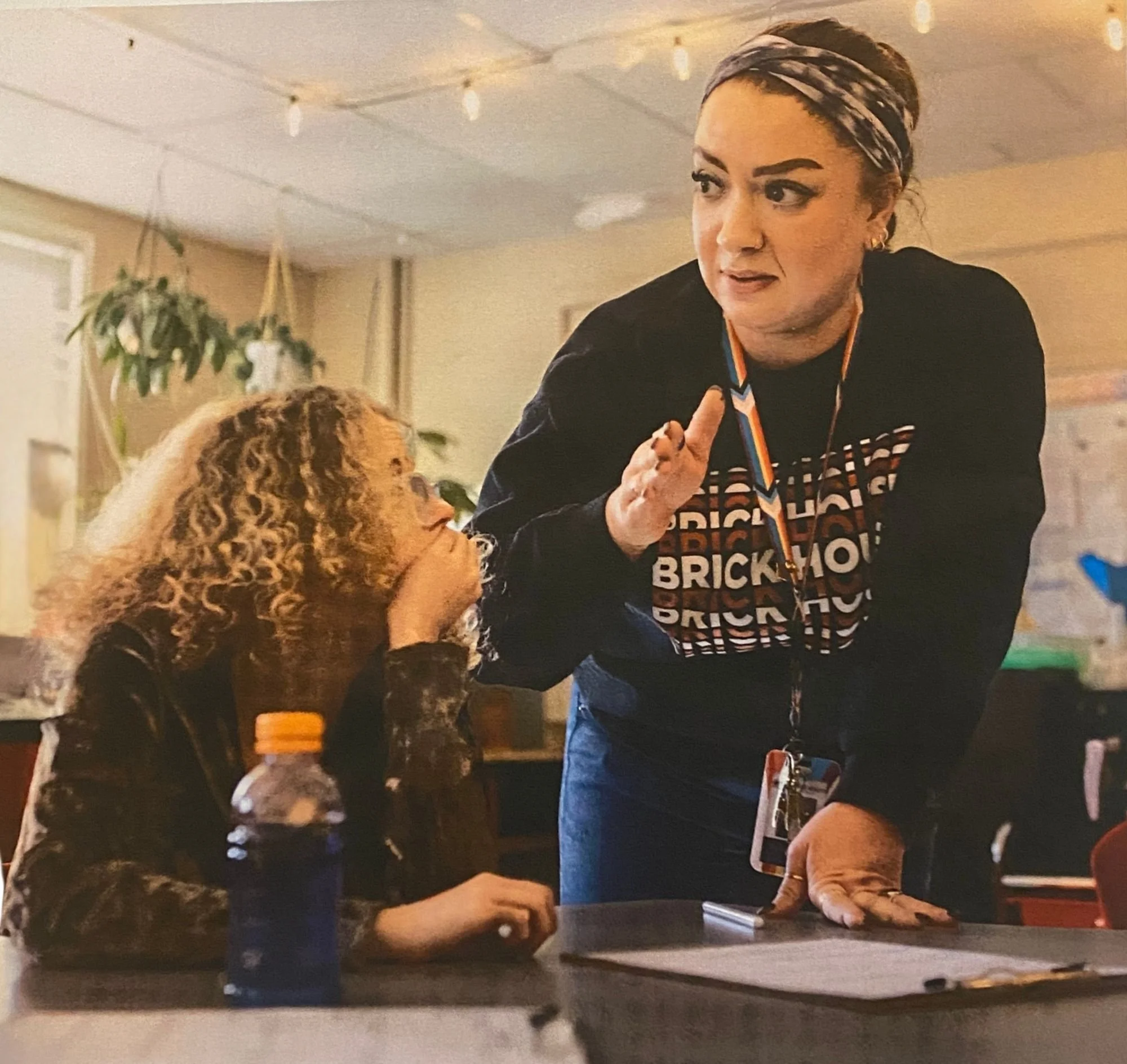 A woman with dark hair tied with a colorful headscarf speaks to a seated woman with curly hair, in a warmly lit indoor setting with hanging string lights and a plant in the background.