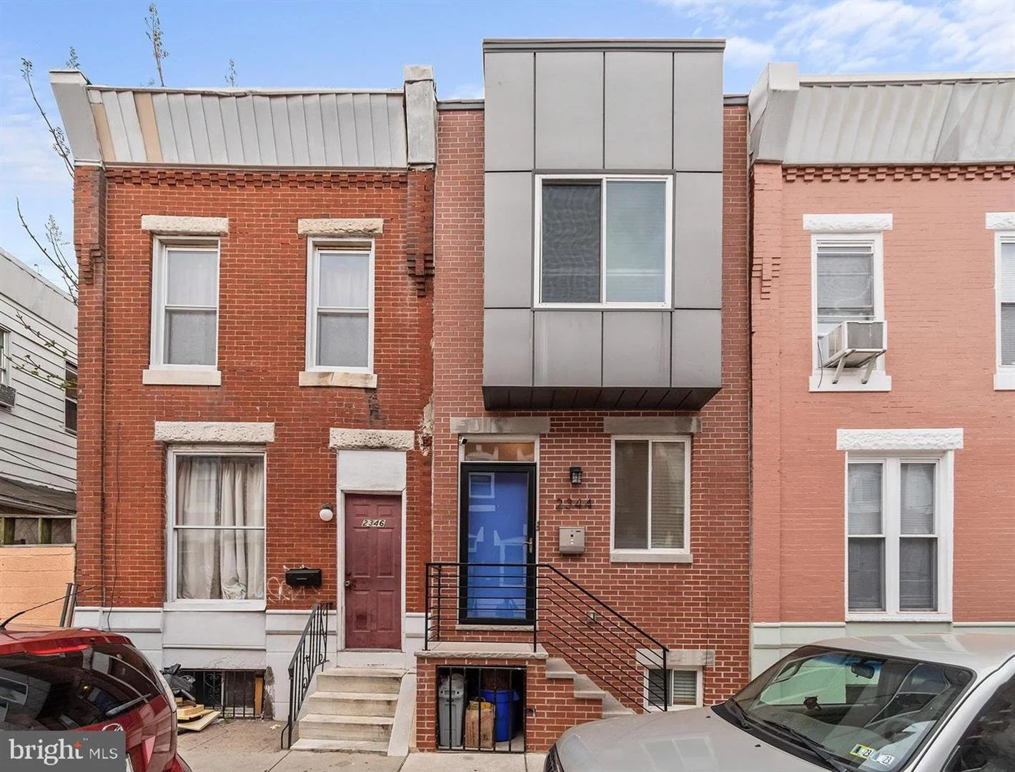 Three row houses in an urban neighborhood. The left house has a brick facade with white window trim and a red door. The middle house, also brick, has a modern metal facade extension over a window and a black door with a ramp. The right house has a brick facade with white window trim and an air conditioning unit in the window. Cars are parked in front of the houses.