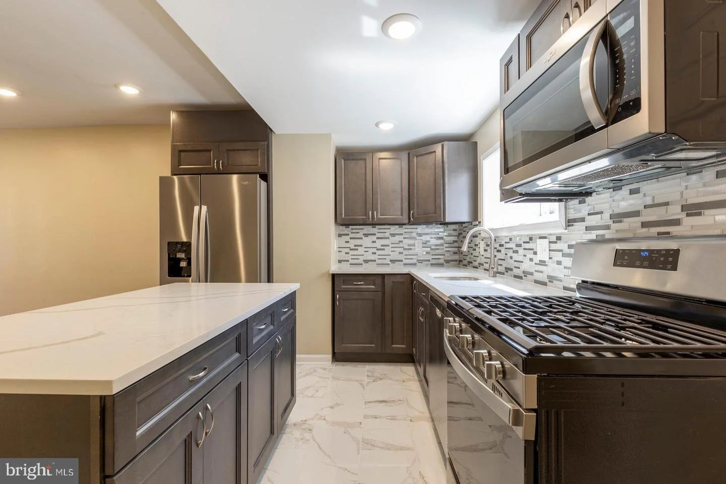 Modern kitchen with gray cabinets, stainless steel appliances, and a white marble countertop island. Features a gas stove, microwave, and refrigerator, with a mosaic tile backsplash and a window above the sink.