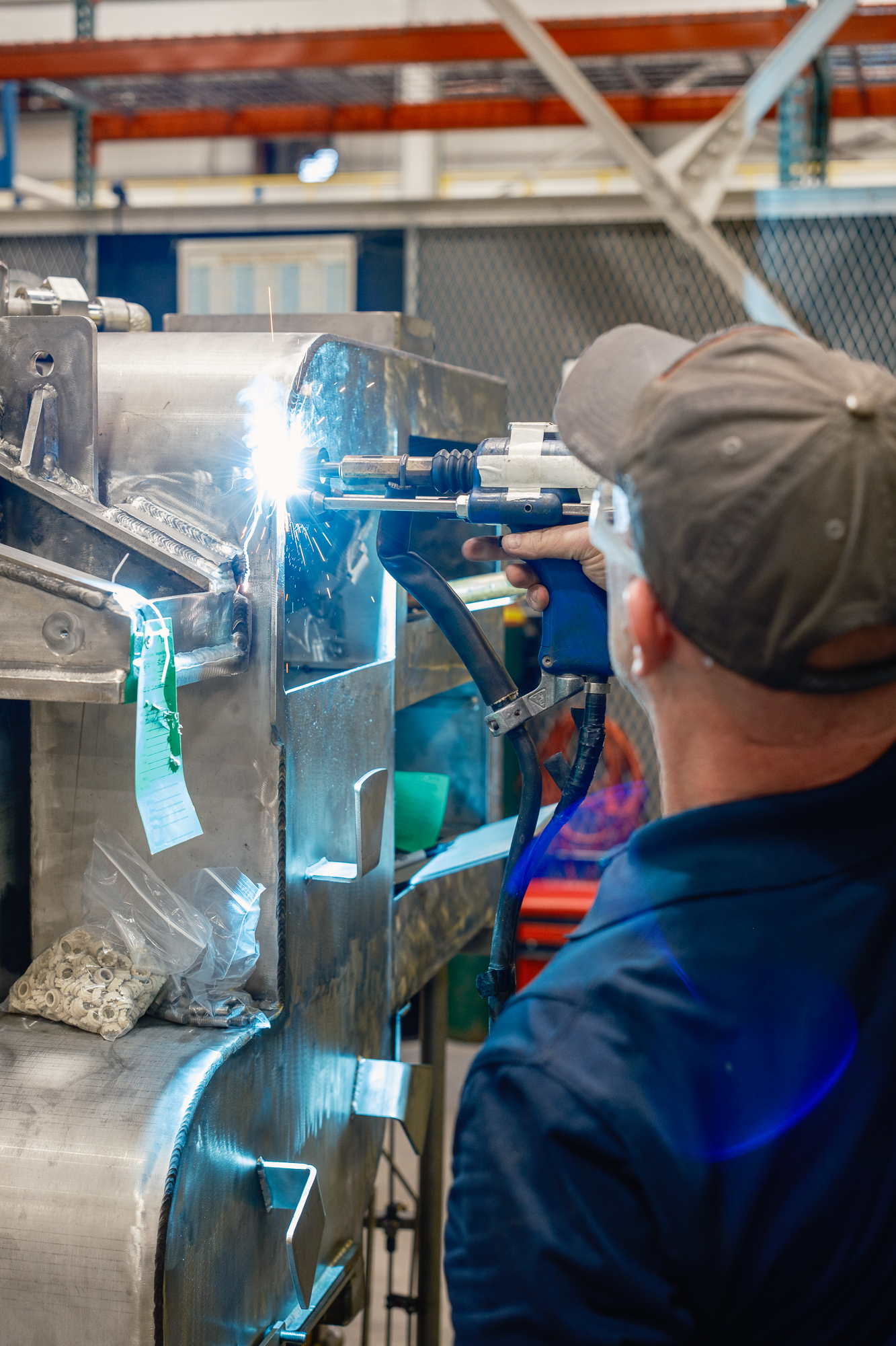A worker in safety glasses and a cap welding a large metal structure inside an industrial workshop. Sparks are visible from the welding process, and there are tools and small parts on a tray beneath the work surface.