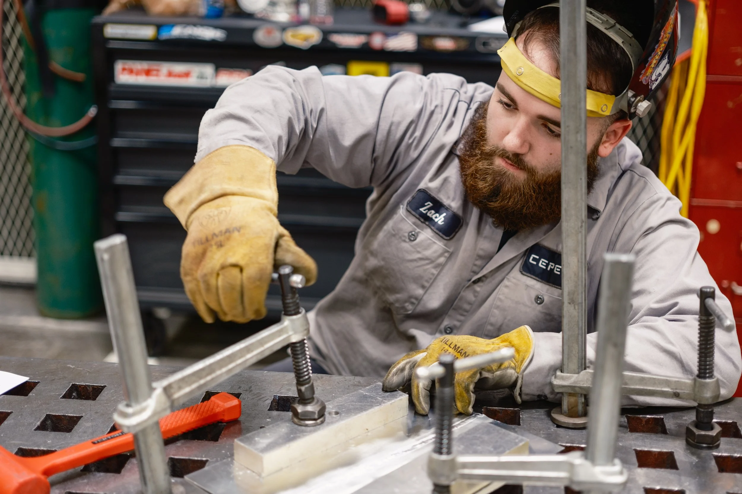 A man wearing a welding helmet and gloves working on a metal frame or structure in a workshop or industrial setting.