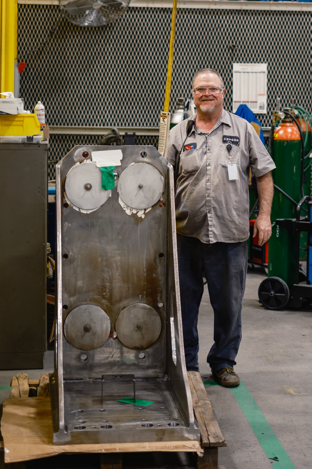 A worker standing in a workshop behind a metal structure with four round openings, on a wooden pallet. The worker is smiling, wearing glasses, a gray work shirt with a name tag, and dark pants. There are tools and machinery in the background.