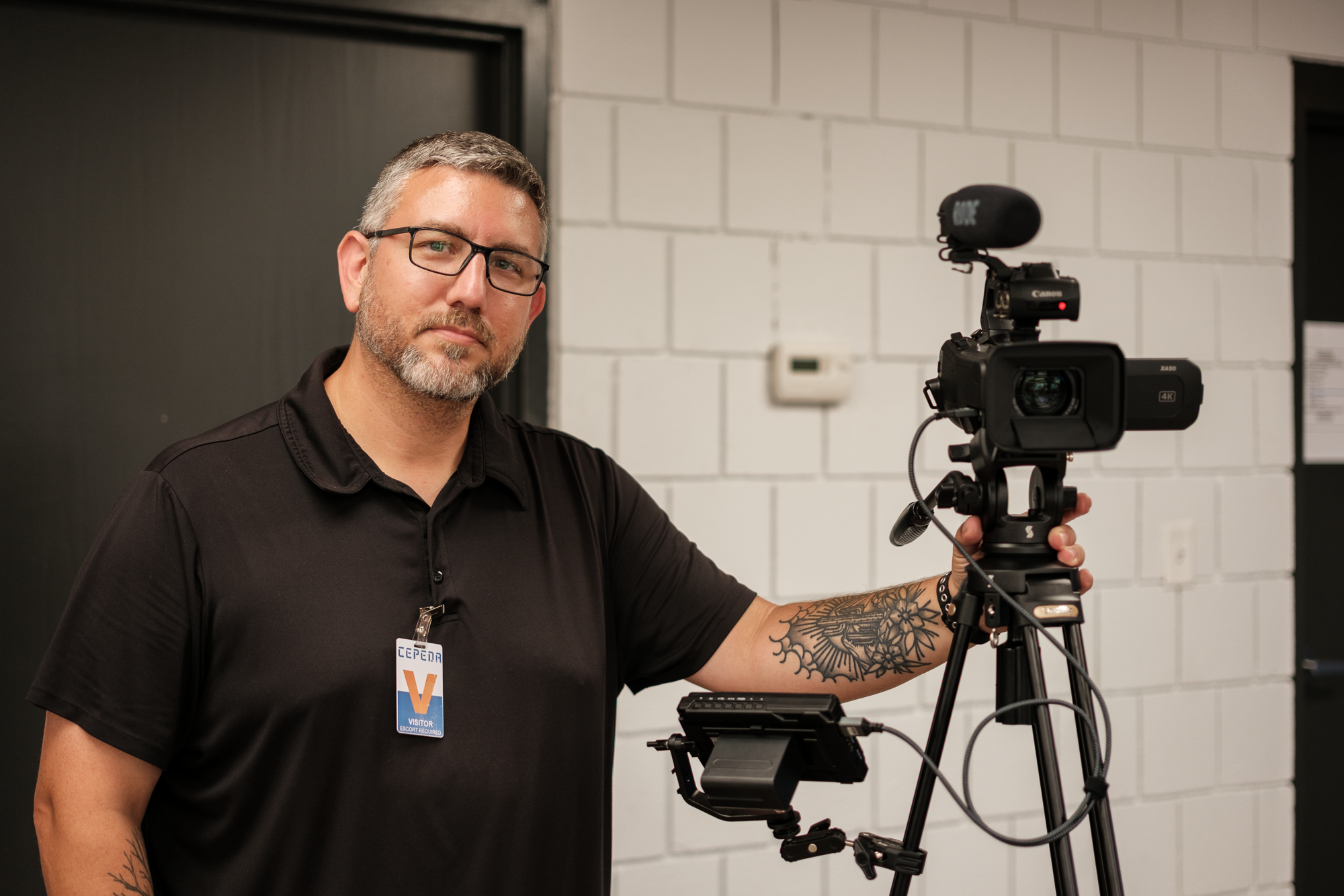 A man in a black shirt with glasses and a beard, operating a professional video camera on a tripod in an indoor setting with white brick walls.